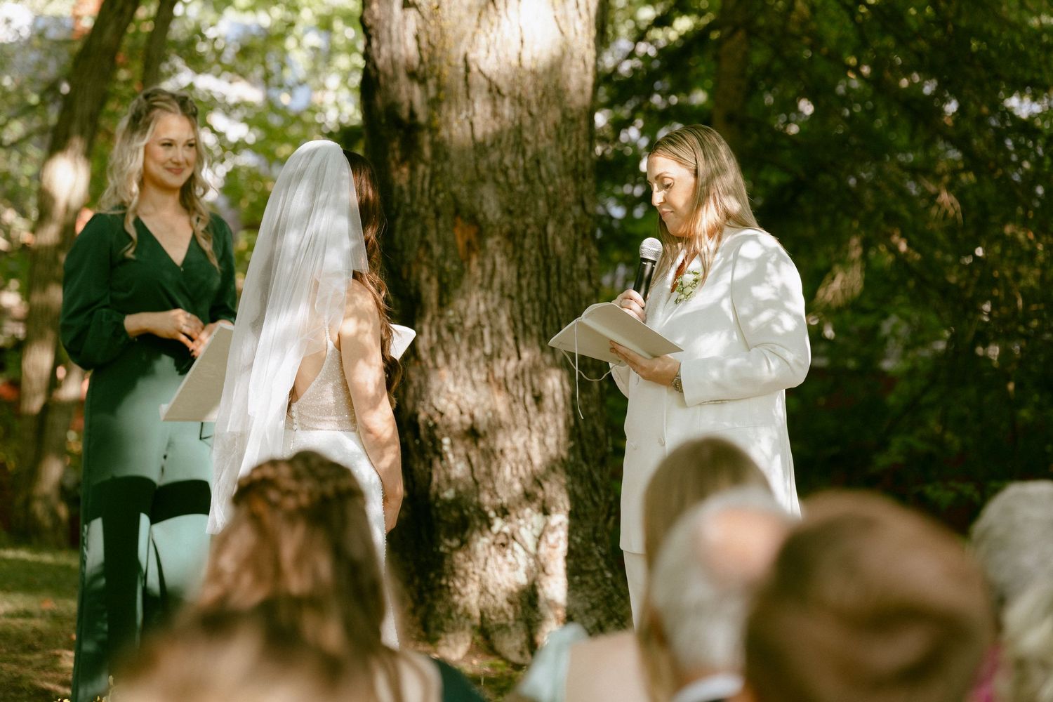 Two brides exchanging vows under a large tree at an outdoor wedding ceremony.