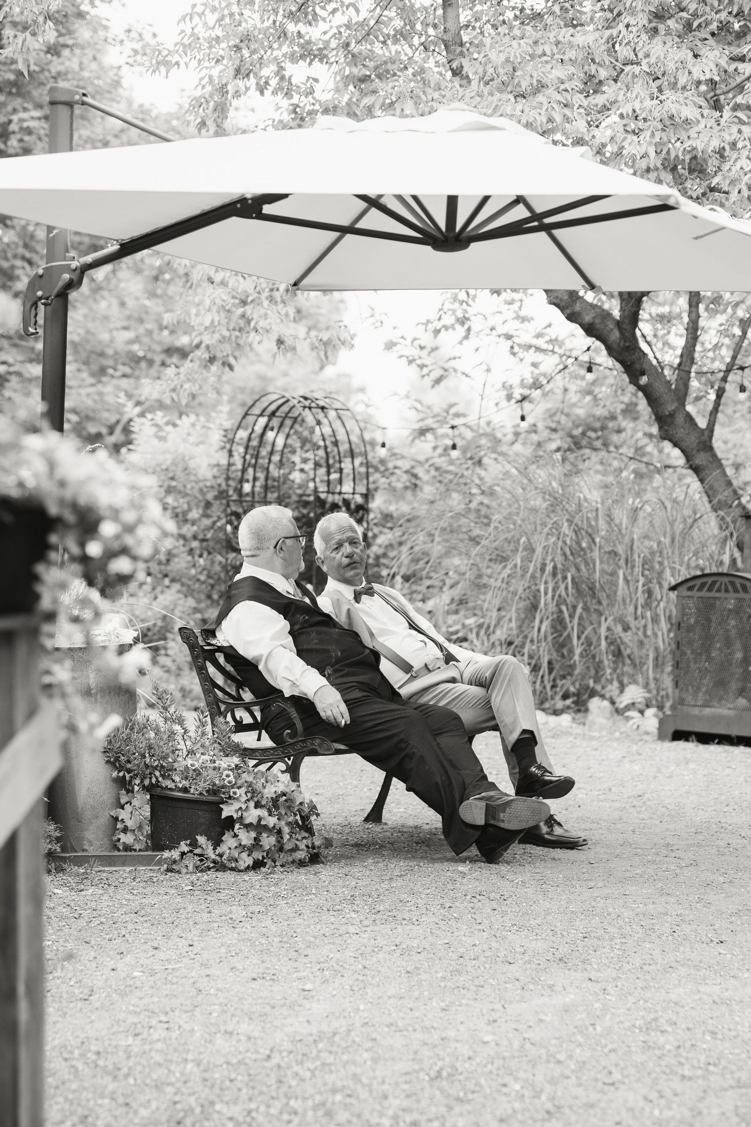 Black and white photo of two elderly men conversing on bench under patio umbrella outdoors.