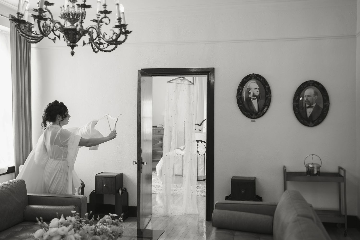 Bride in white dress dancing in elegant room with portrait paintings and chandelier, black and white photo.