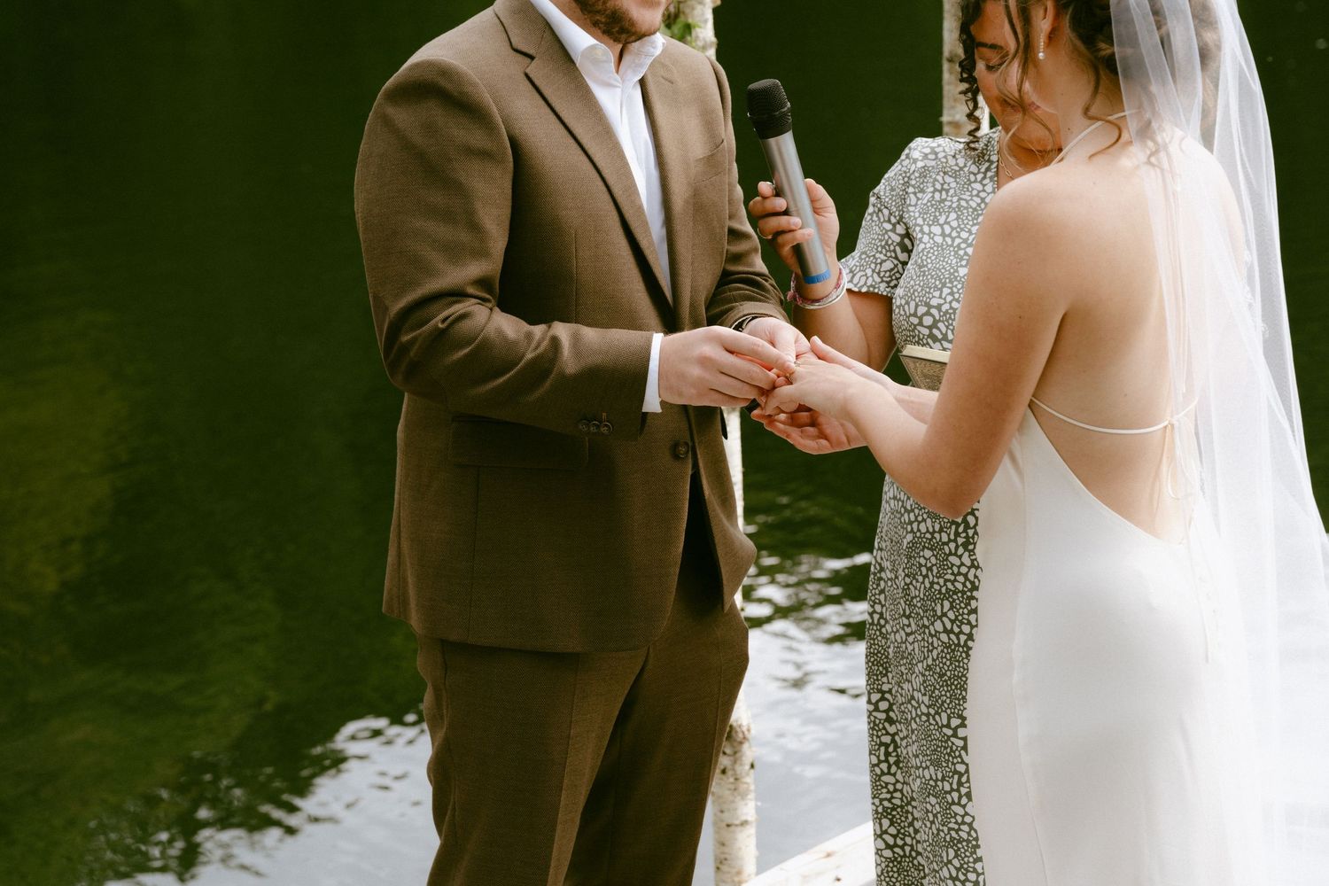 Bride places wedding ring on grooms finger during lakeside dock ceremony with officiant nearby.