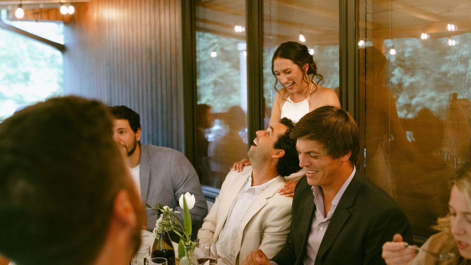 Bride laughing with wedding guests seated at a reception dinner table in a warmly lit venue.