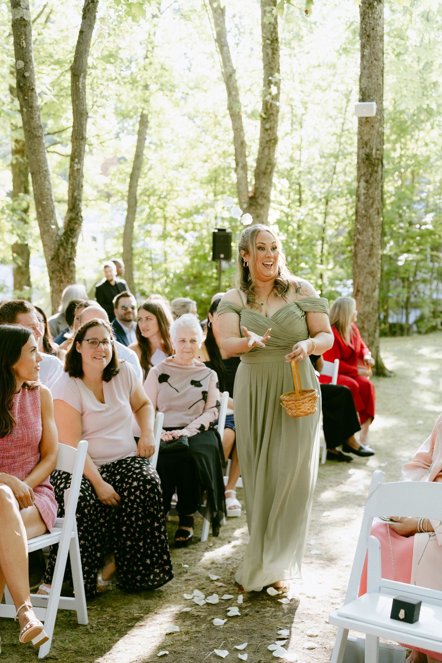 Smiling bridesmaid in sage green gown carries flower basket down outdoor wedding ceremony aisle.