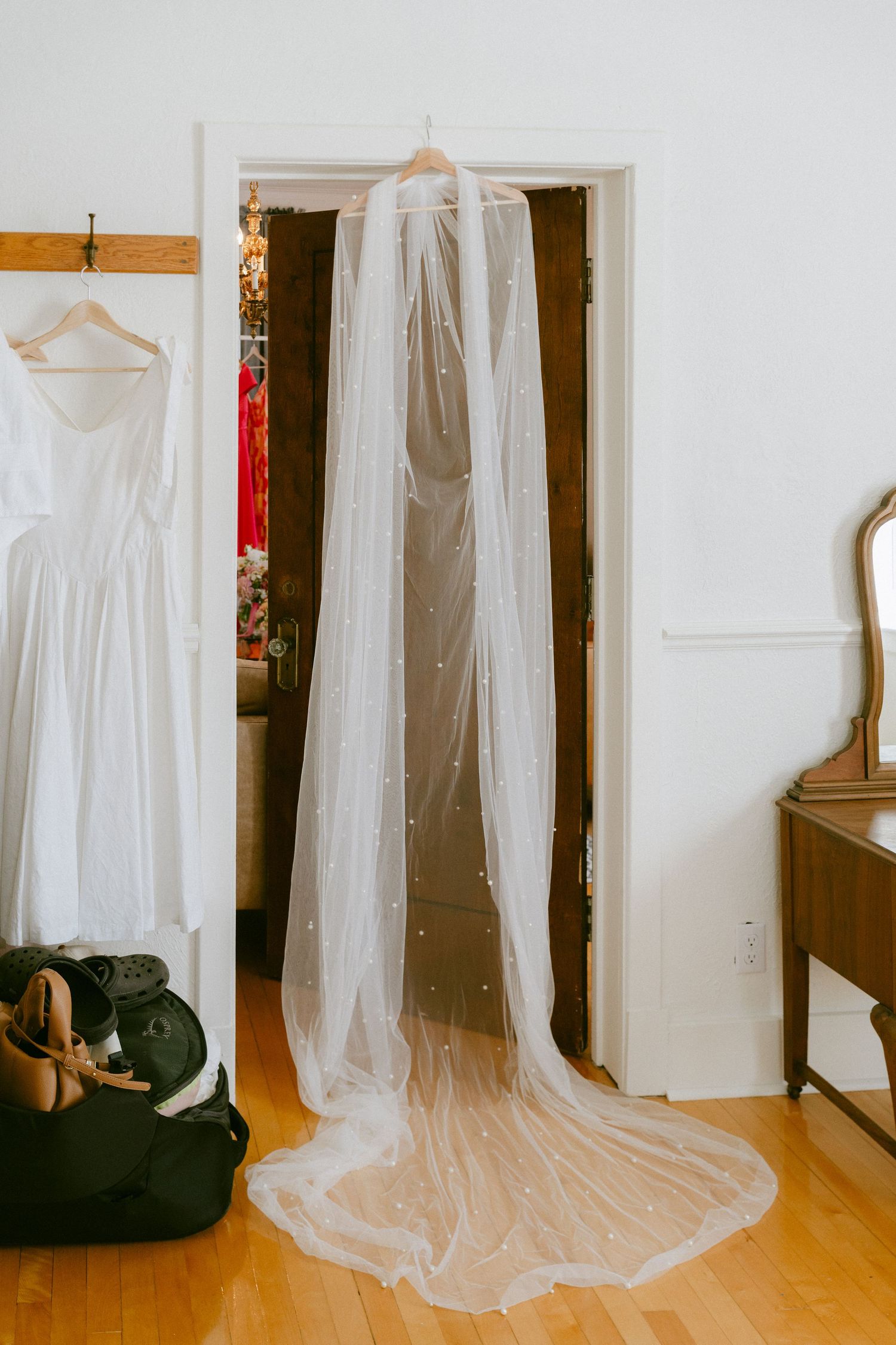 Long pearl-embellished wedding veil hanging in a doorway beside a white bridal gown.