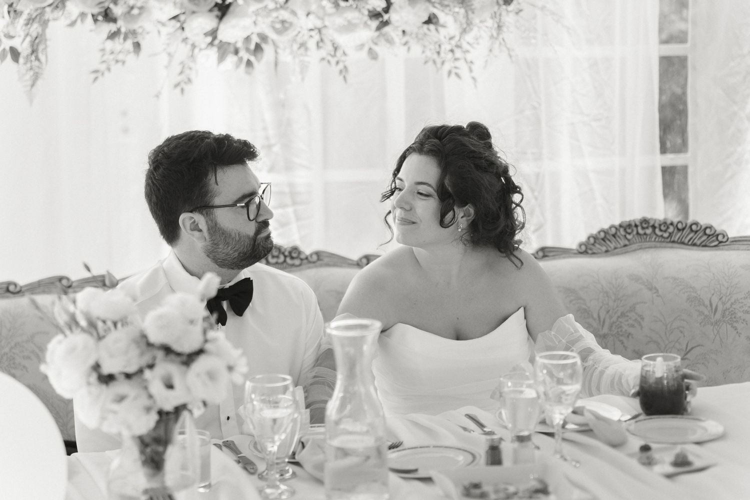 Black and white photo of bride and groom smiling at each other at wedding reception sweetheart table.