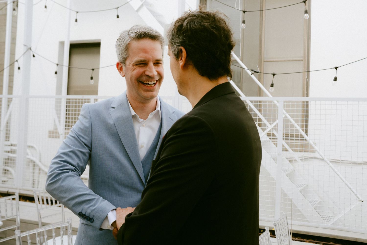 Two men in suits laughing and shaking hands at outdoor wedding reception with string lights.