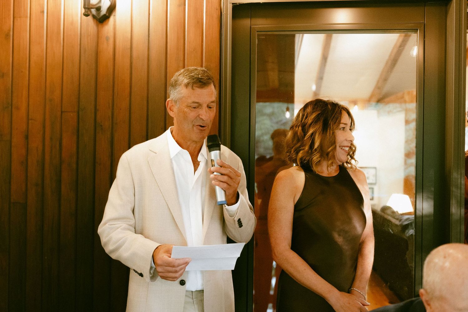 Man in white suit giving a speech at a wedding reception while a woman in black dress smiles beside him.