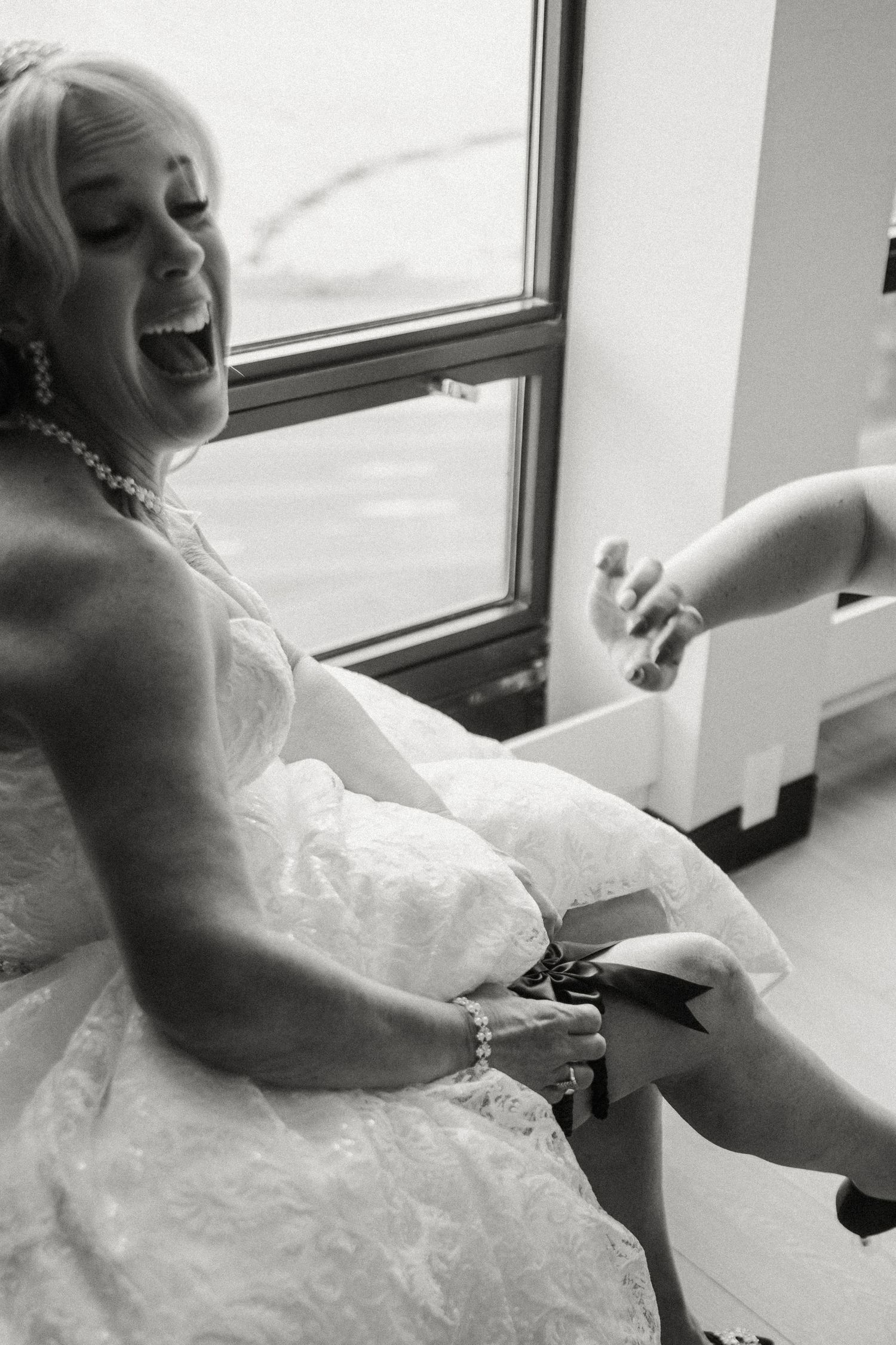 Laughing bride in lace wedding gown having shoes put on during joyful bridal preparations.