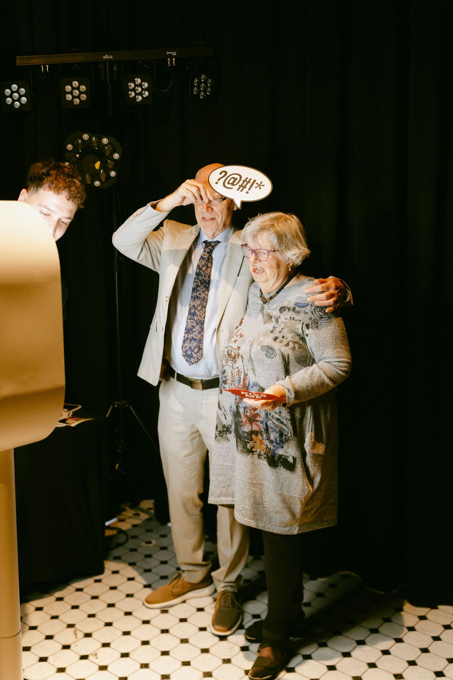 Couple posing at wedding photo booth with fun speech bubble prop on checkered floor.