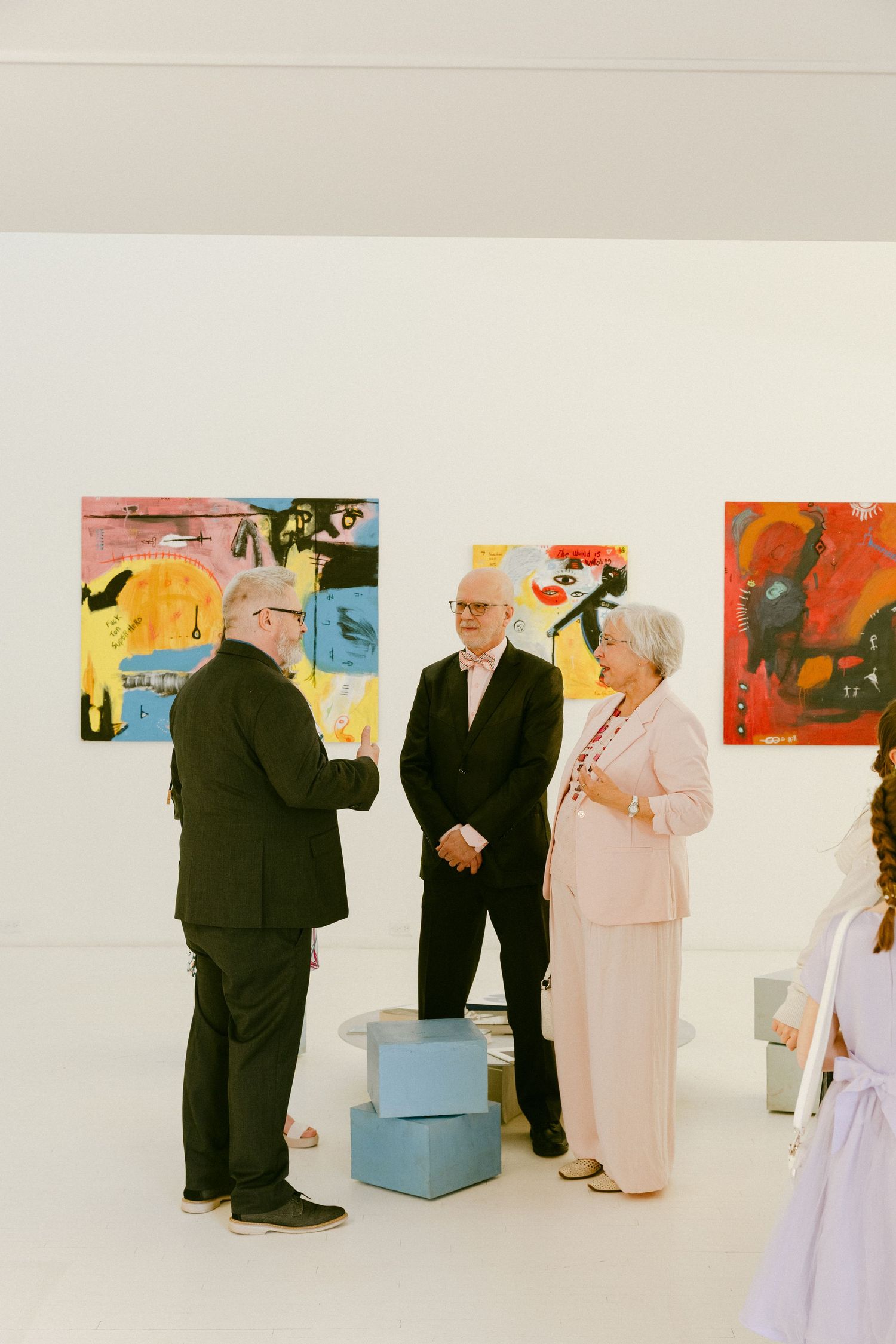 Three elegantly dressed guests conversing in a modern white art gallery with colorful abstract paintings on the walls.