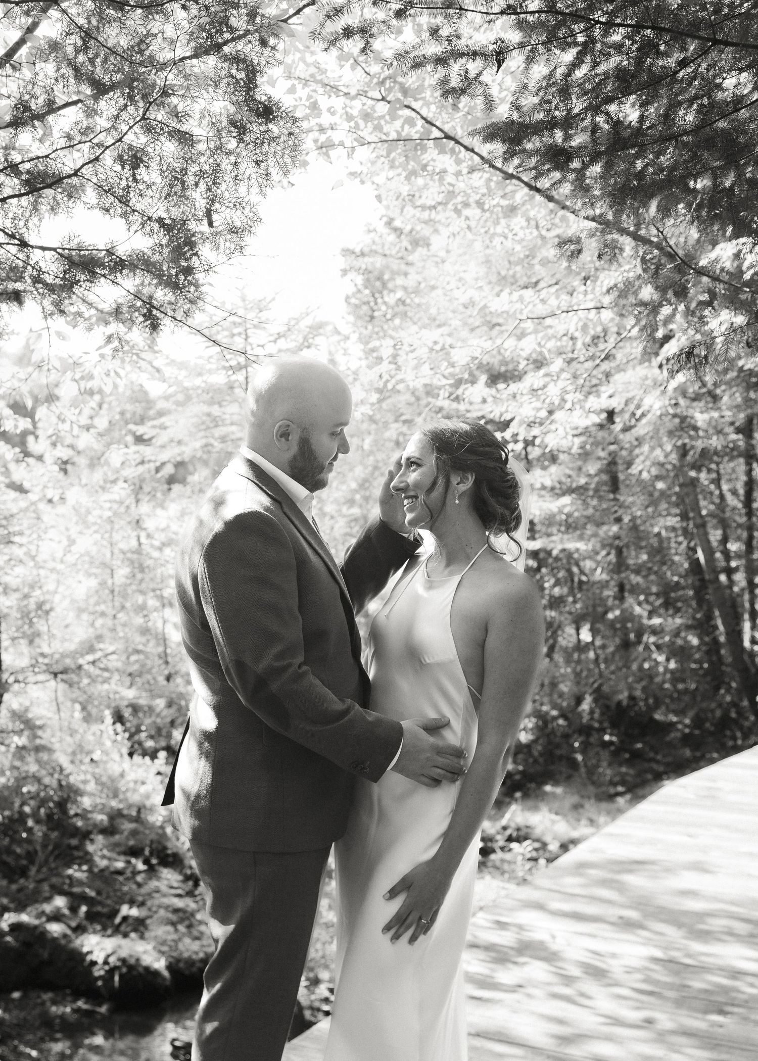 Black and white portrait of smiling bride and groom embracing under sunlit trees in a forest setting.