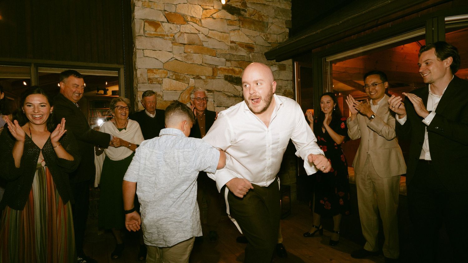 Groom in white shirt dancing energetically with young boy at wedding reception.