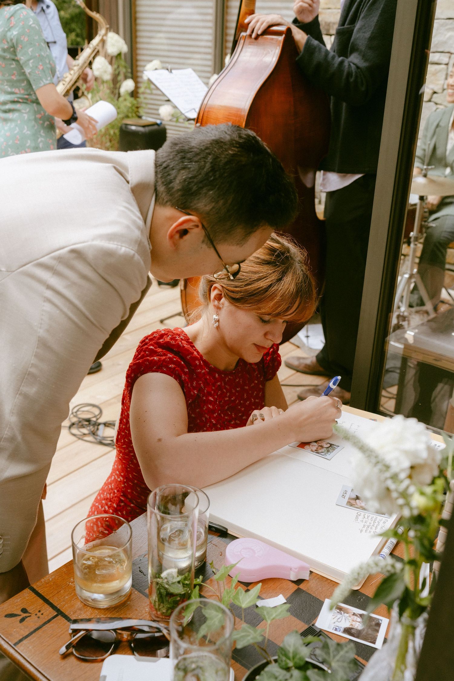 Woman in red dress signing wedding guest book at table with drinks and greenery while man leans over.