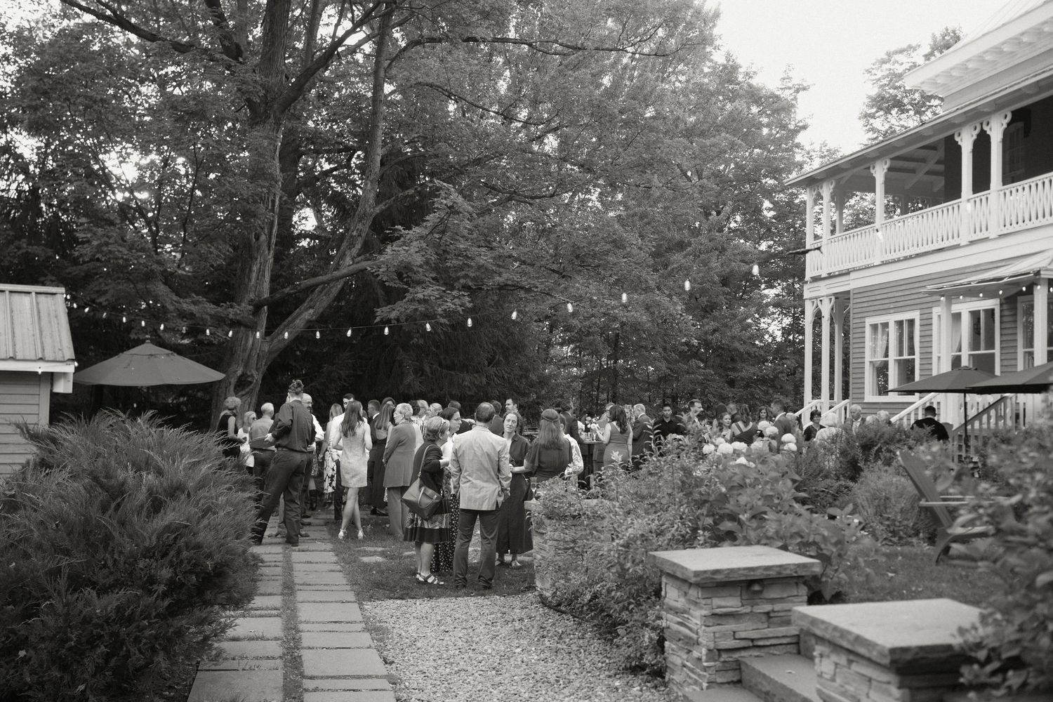 Wedding guests mingle in a lush garden beside an elegant historic home with string lights at dusk.