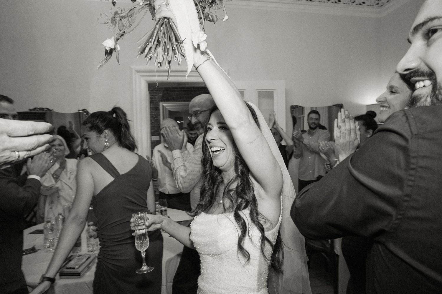 Bride in white gown dancing joyfully with raised arm among celebrating wedding guests.