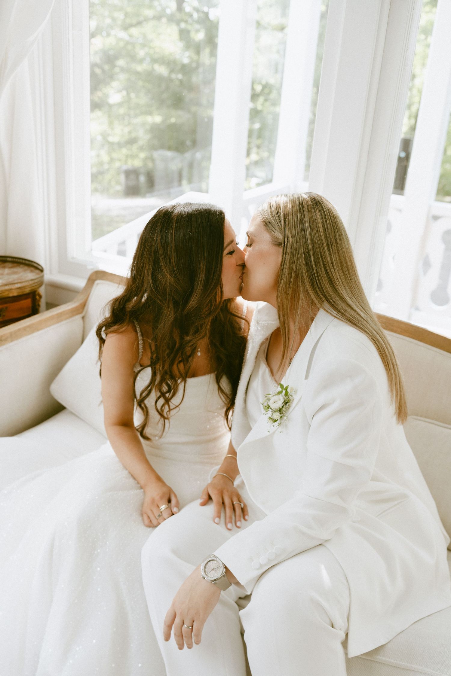 Two brides share a kiss while seated on a sofa near bright windows.