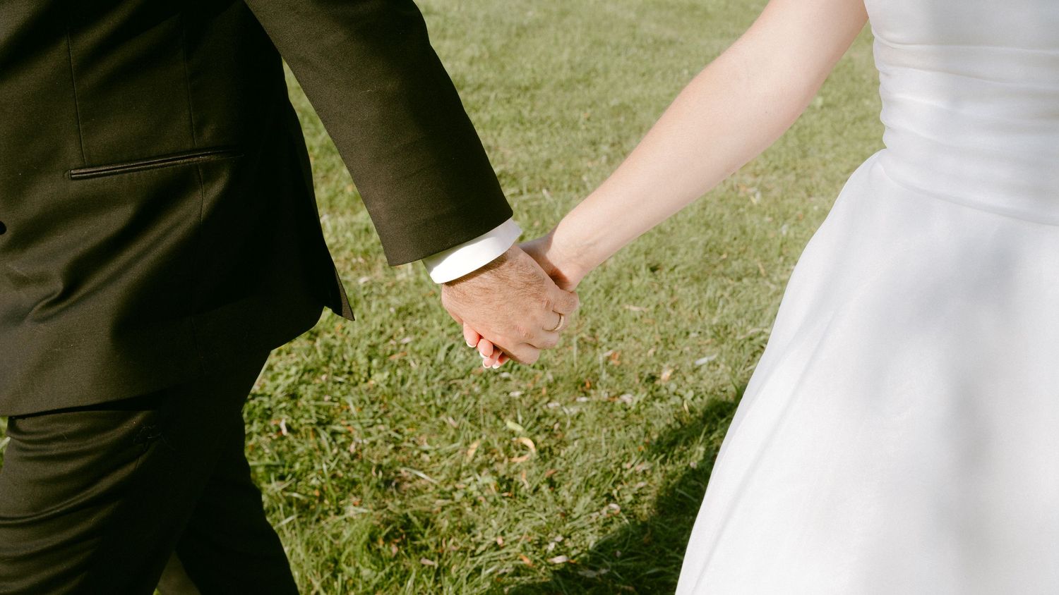 Close-up of bride and groom holding hands on green grass at an outdoor wedding ceremony.