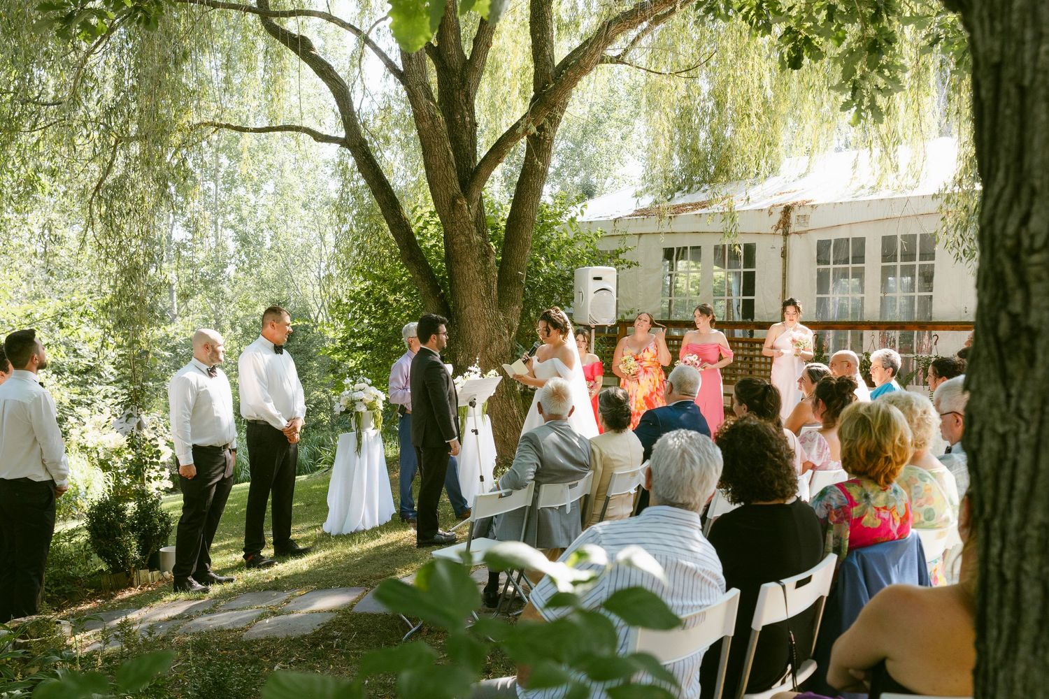 Wedding ceremony under willow tree with guests seated on white chairs outdoors.