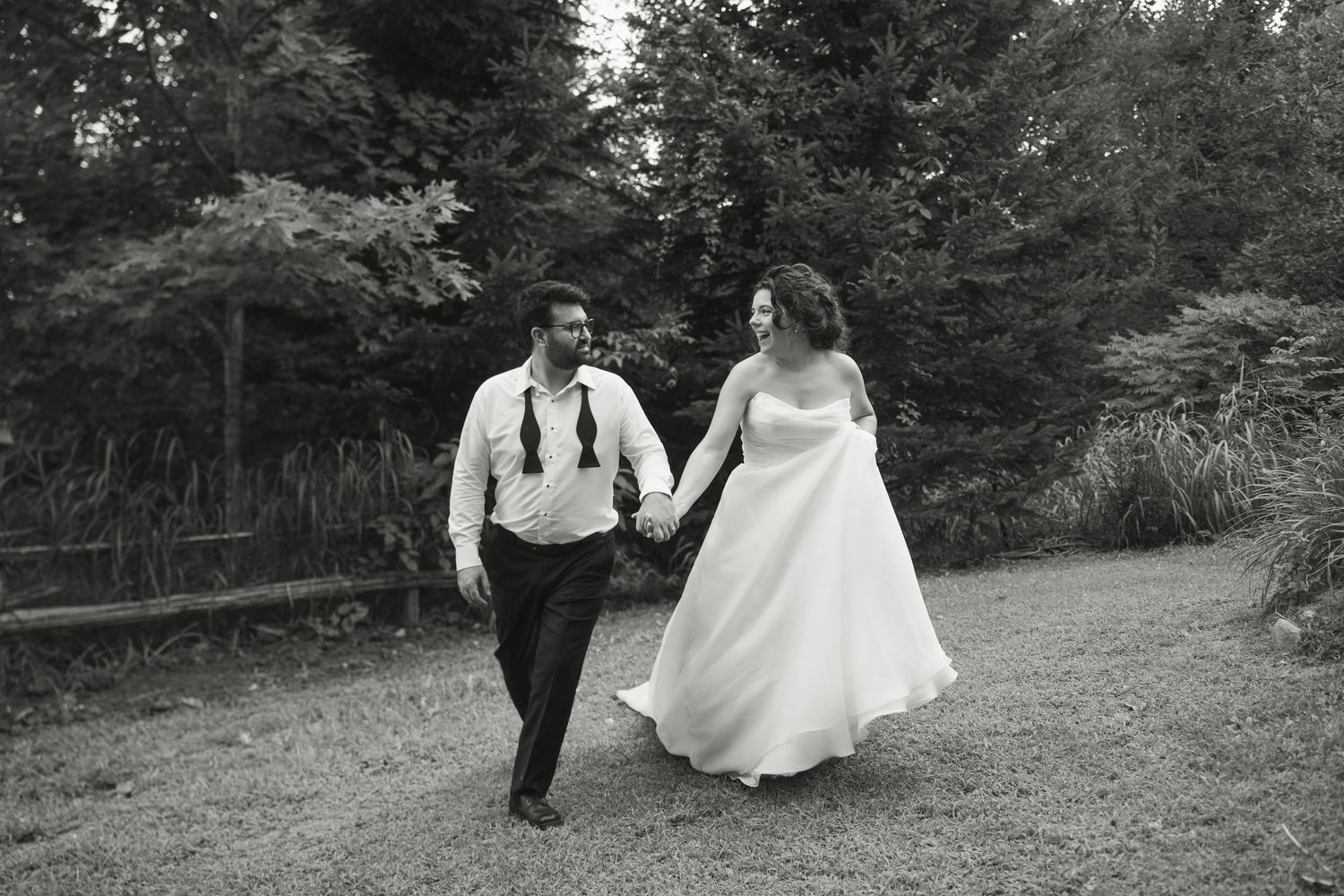 Black and white photo of bride and groom joyfully walking hand in hand through a wooded path.