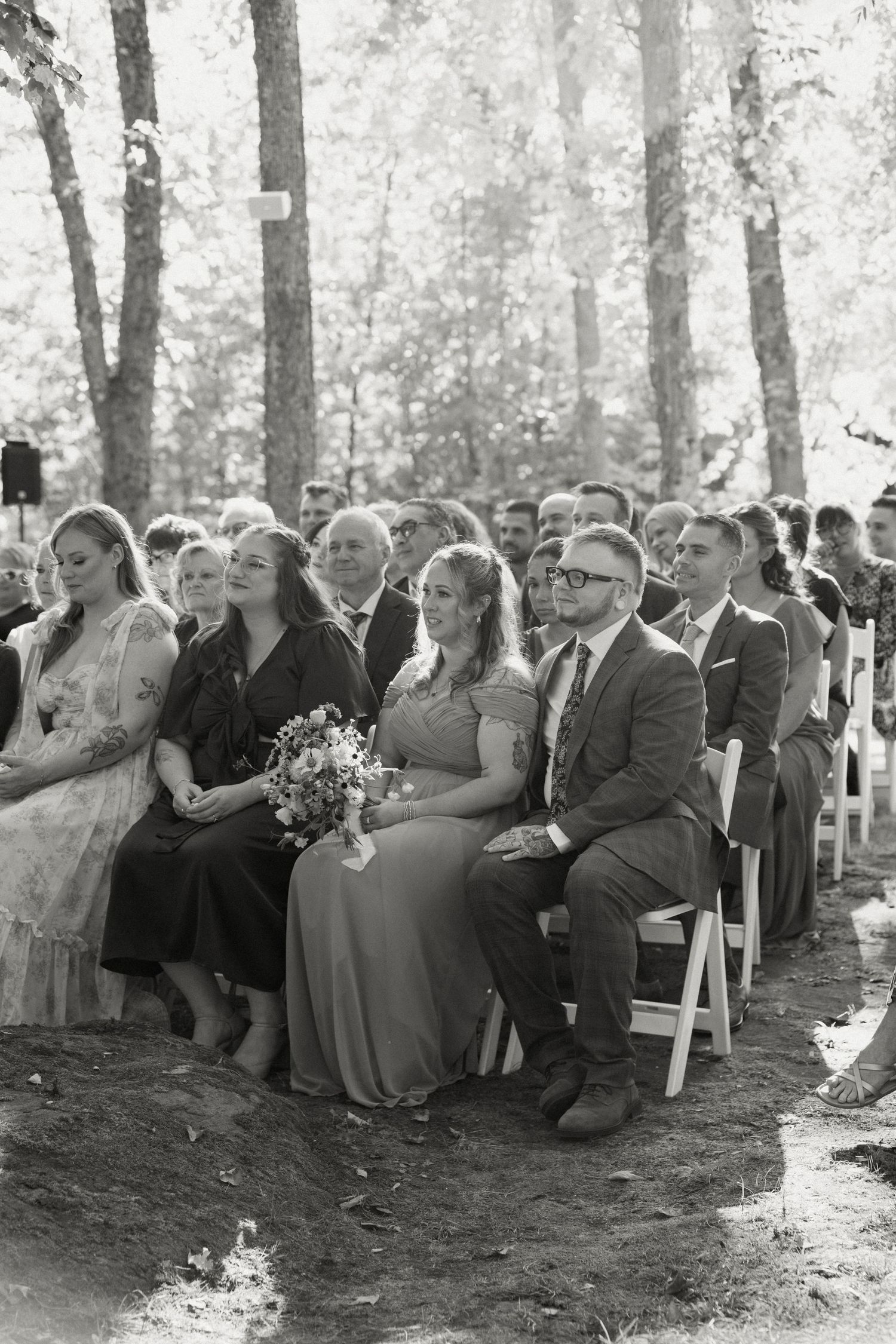 Black and white photo of wedding guests seated outdoors watching a forest ceremony.