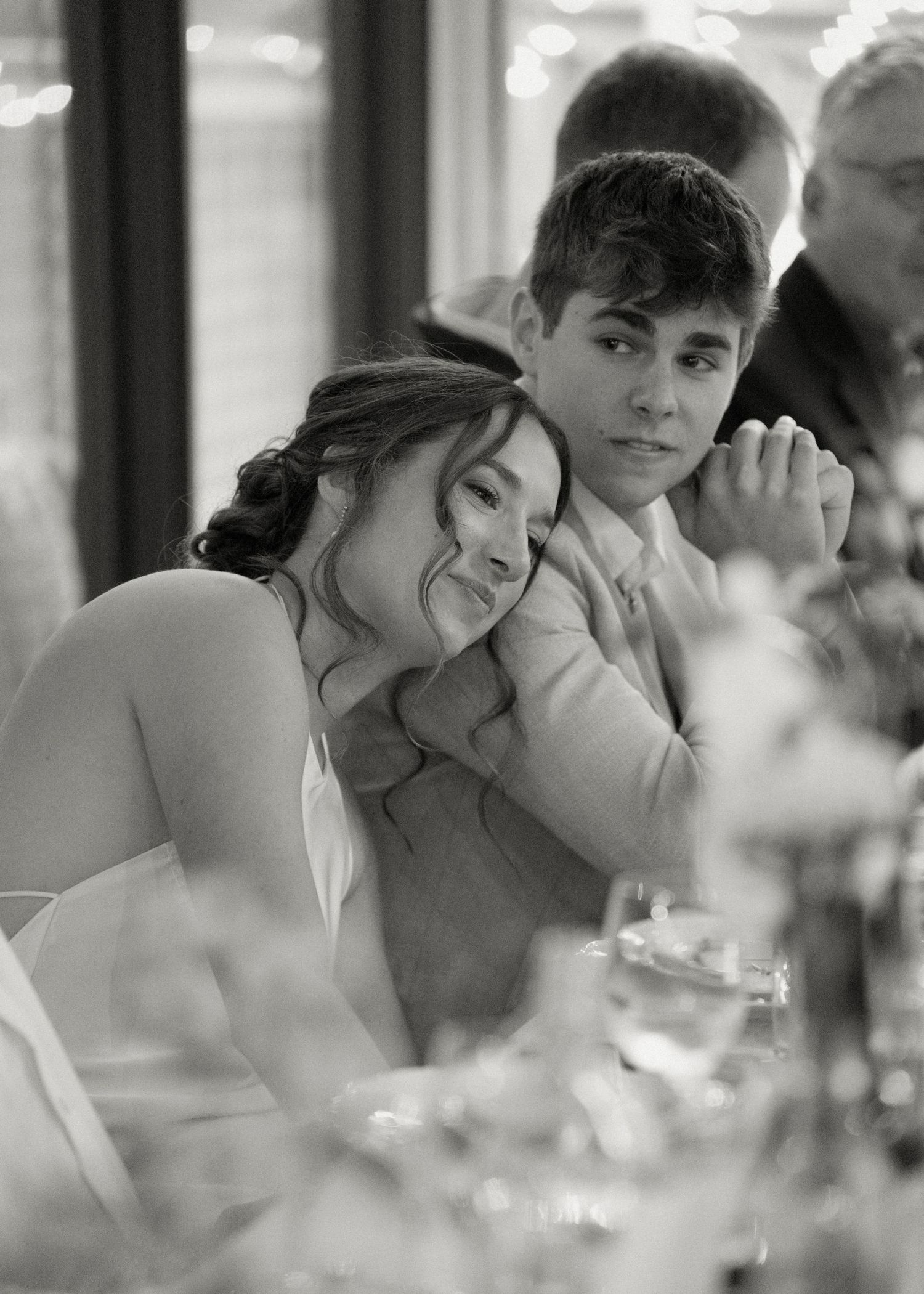 Black and white photo of a young woman resting her head on someone's shoulder at a wedding reception.