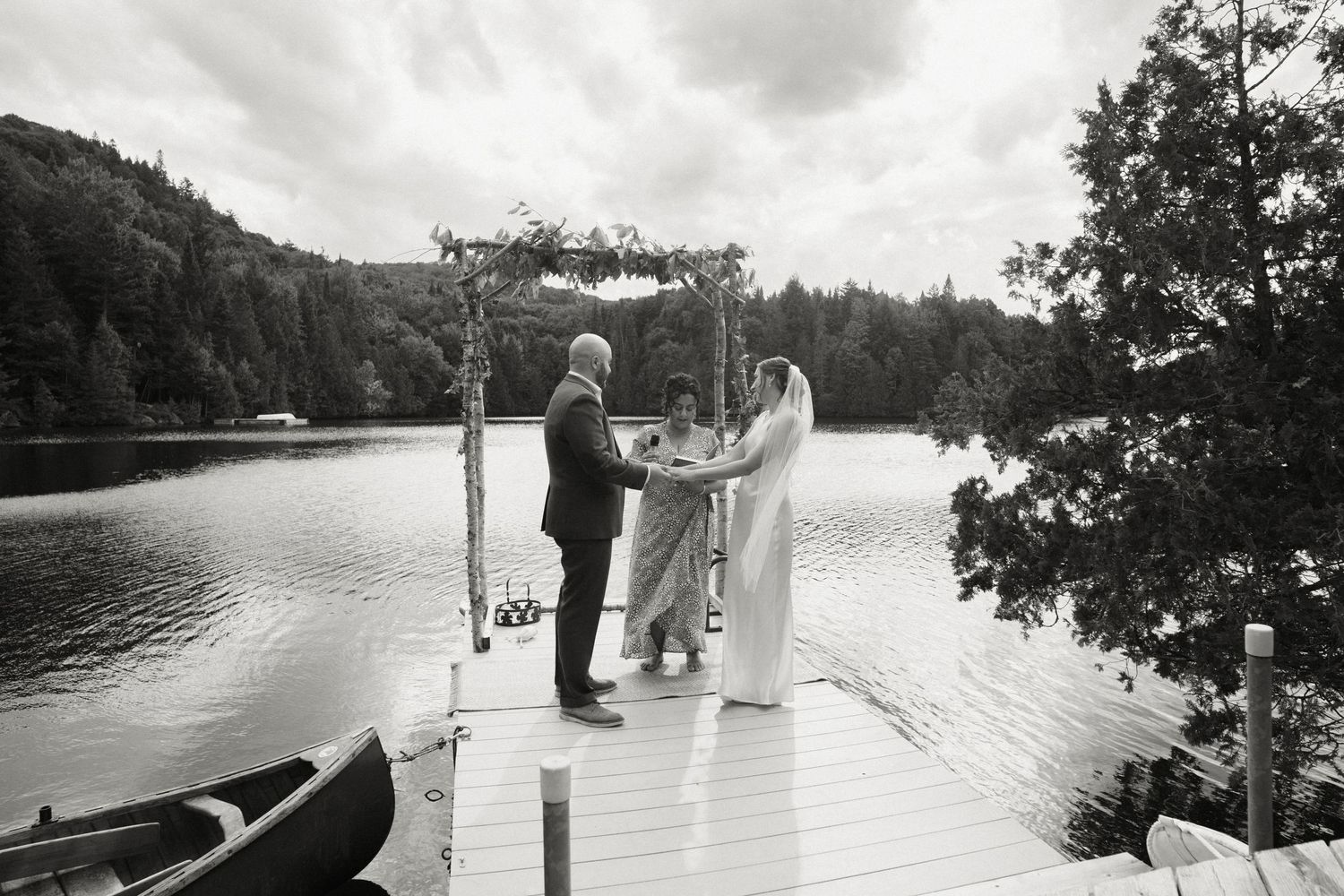 Couple exchanges vows at a decorated arch on a wooden dock with a serene lake backdrop.