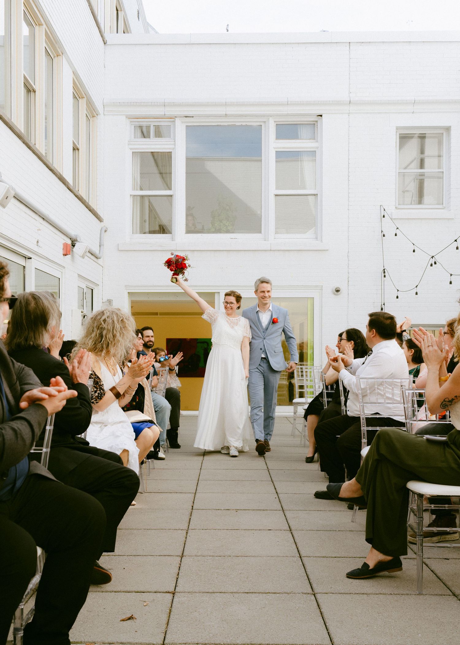 Newlywed couple joyfully walking down the aisle as guests applaud in a bright outdoor courtyard ceremony.