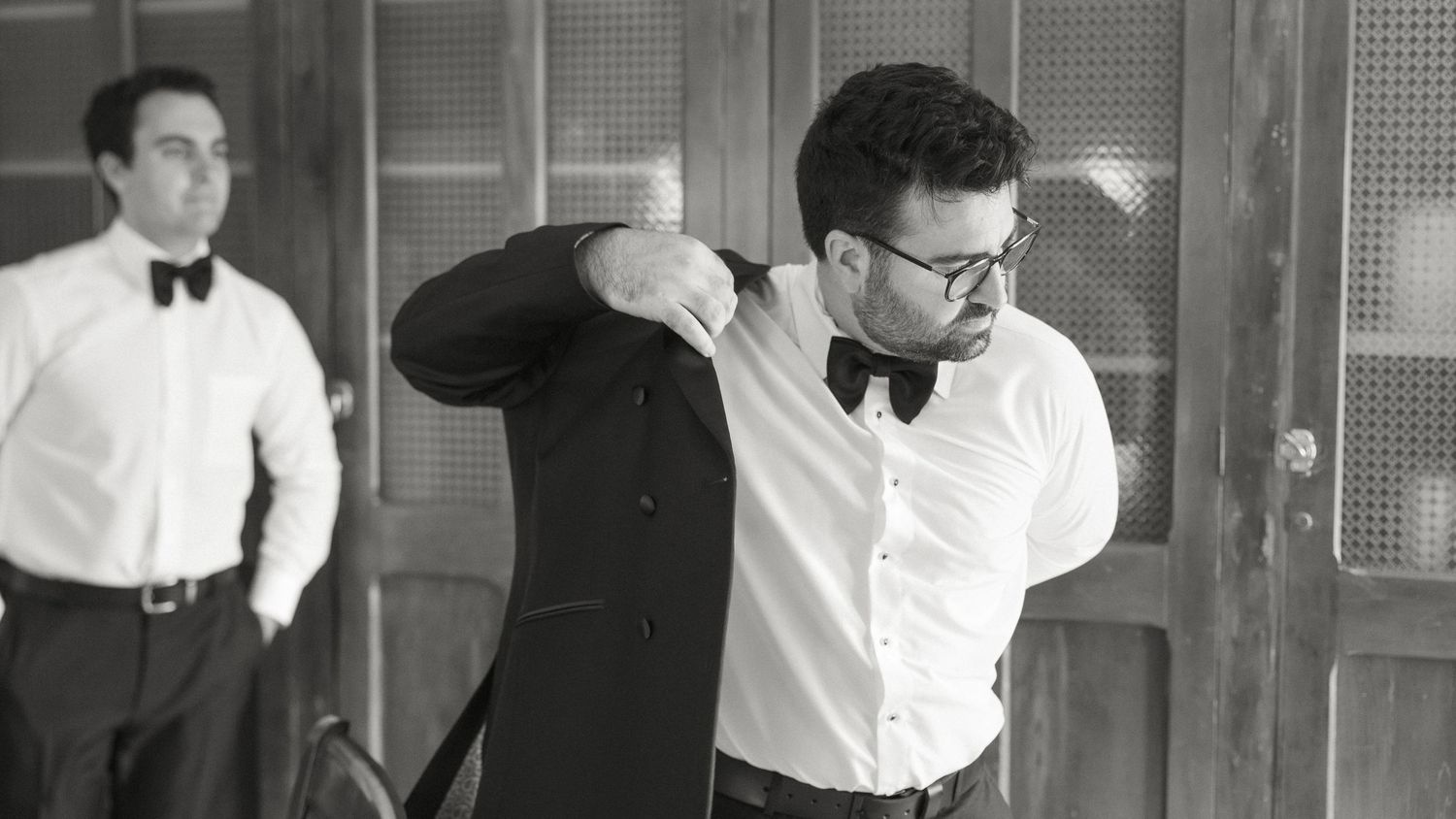 A groom puts on his tuxedo jacket while a groomsman watches in black and white.
