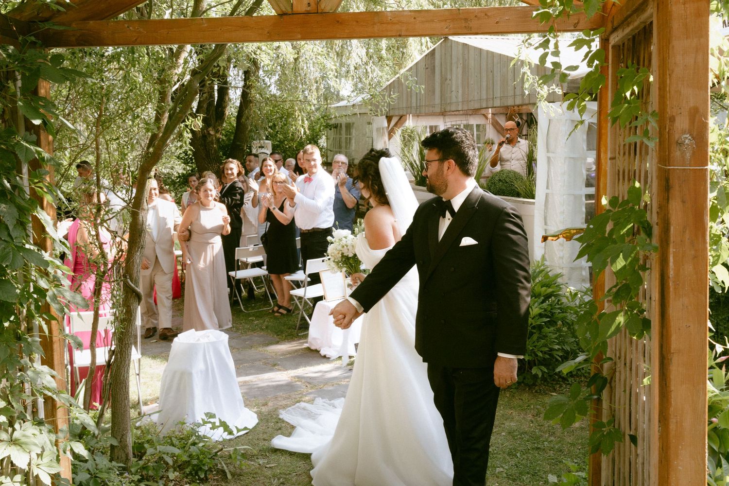 Bride and groom walk hand in hand through a wooden pergola at an outdoor garden wedding ceremony.