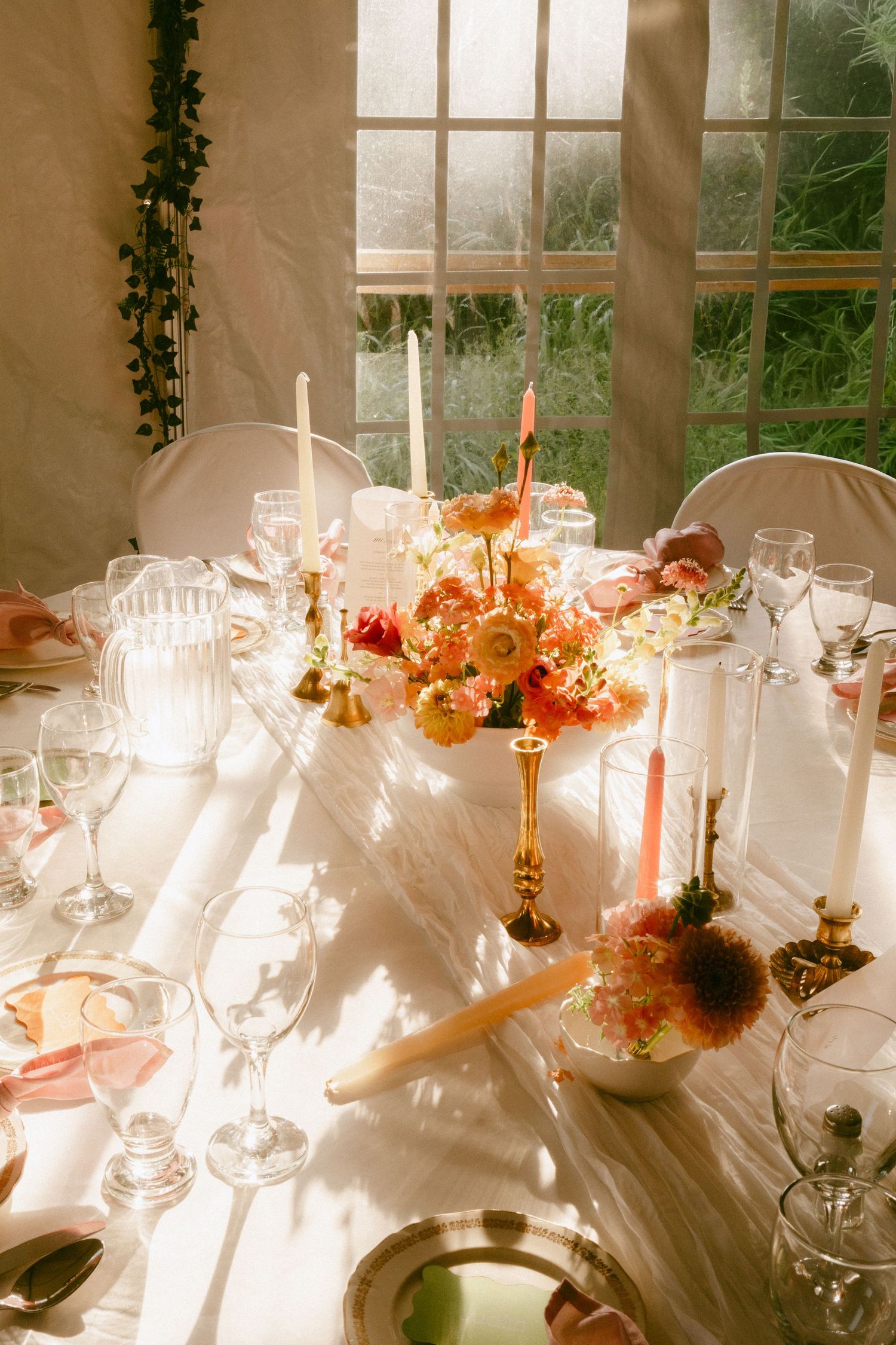 Sunlit wedding reception table with orange and pink floral centerpiece, gold candlesticks, and crystal glasses.