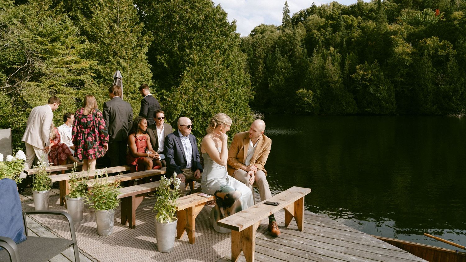 Wedding guests seated on wooden benches by a serene lakeside during an outdoor ceremony.