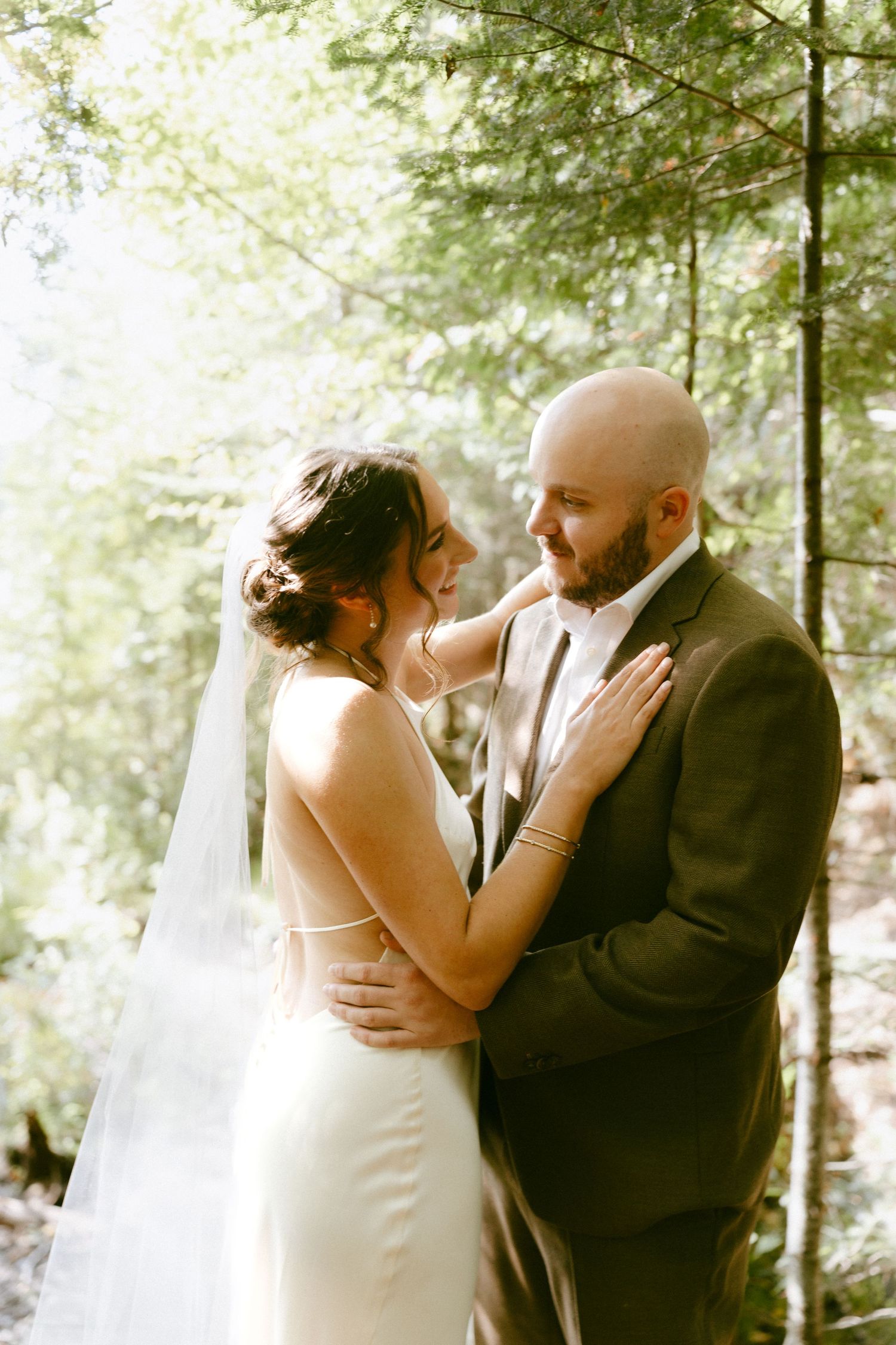 Bride and groom share a tender forehead-to-forehead moment in a sun-drenched forest during their wedding portraits.