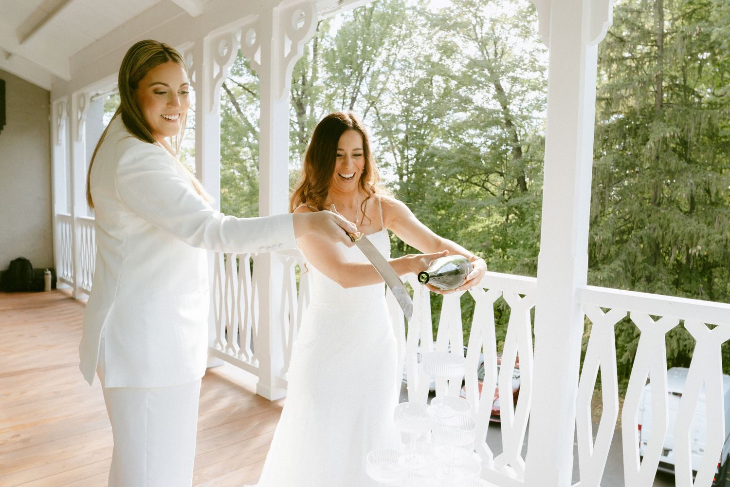 Two brides in white suits and gowns laugh while pouring champagne on a decorated porch.