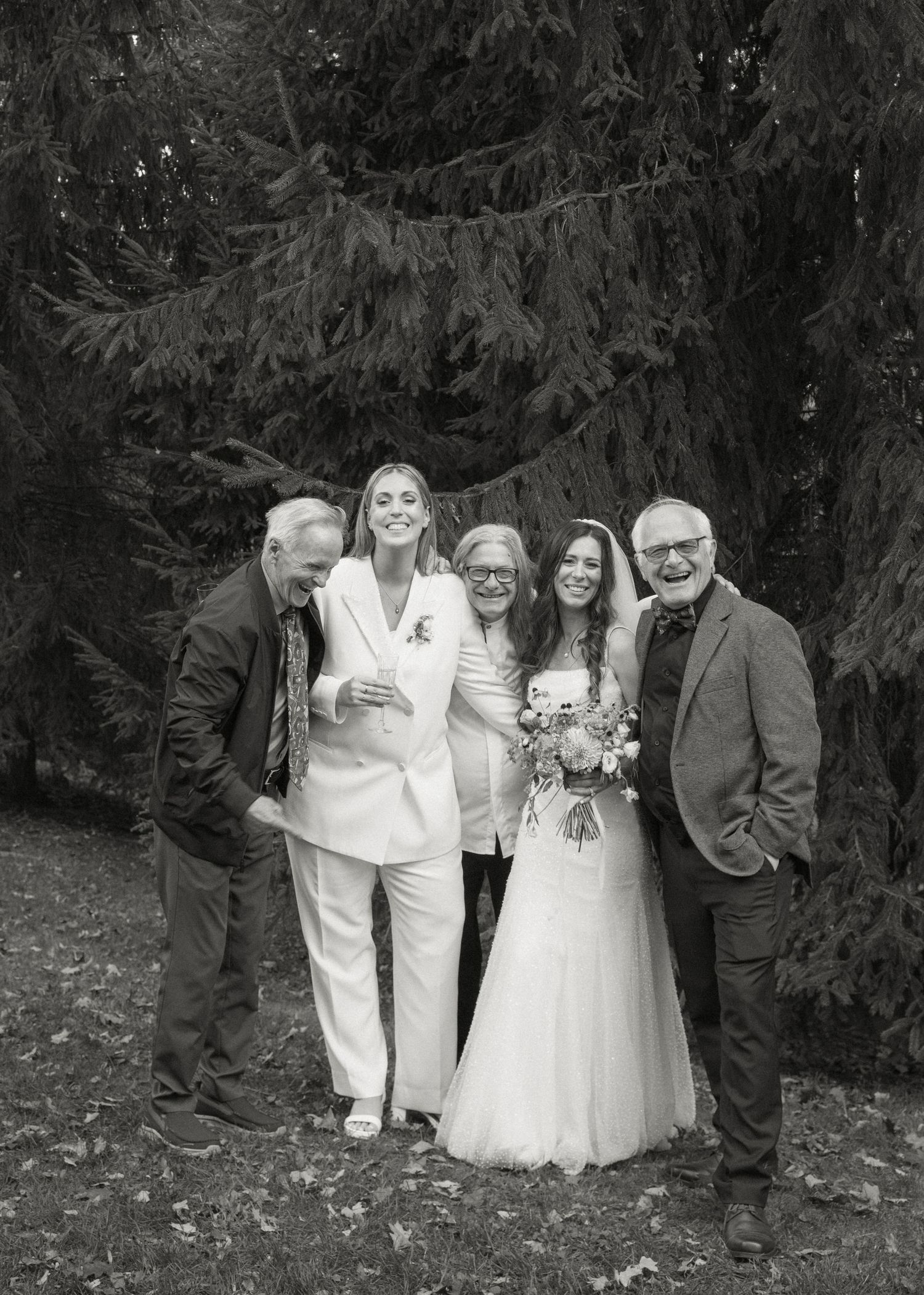 Two brides pose with three family members for a black and white portrait in front of evergreen trees.