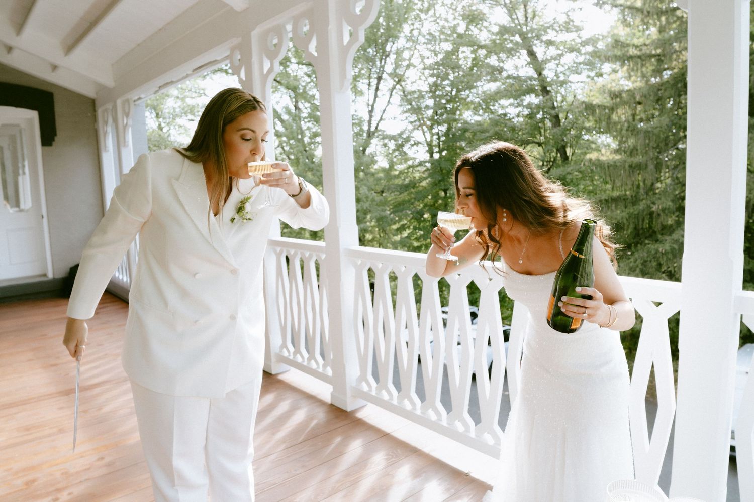 Two brides in white toast with champagne glasses on a sunlit porch surrounded by trees.