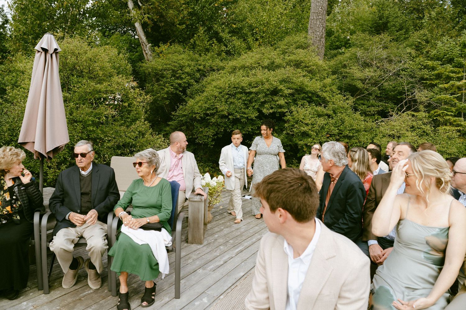 Outdoor wedding ceremony on a wooden deck with guests seated among lush green trees.