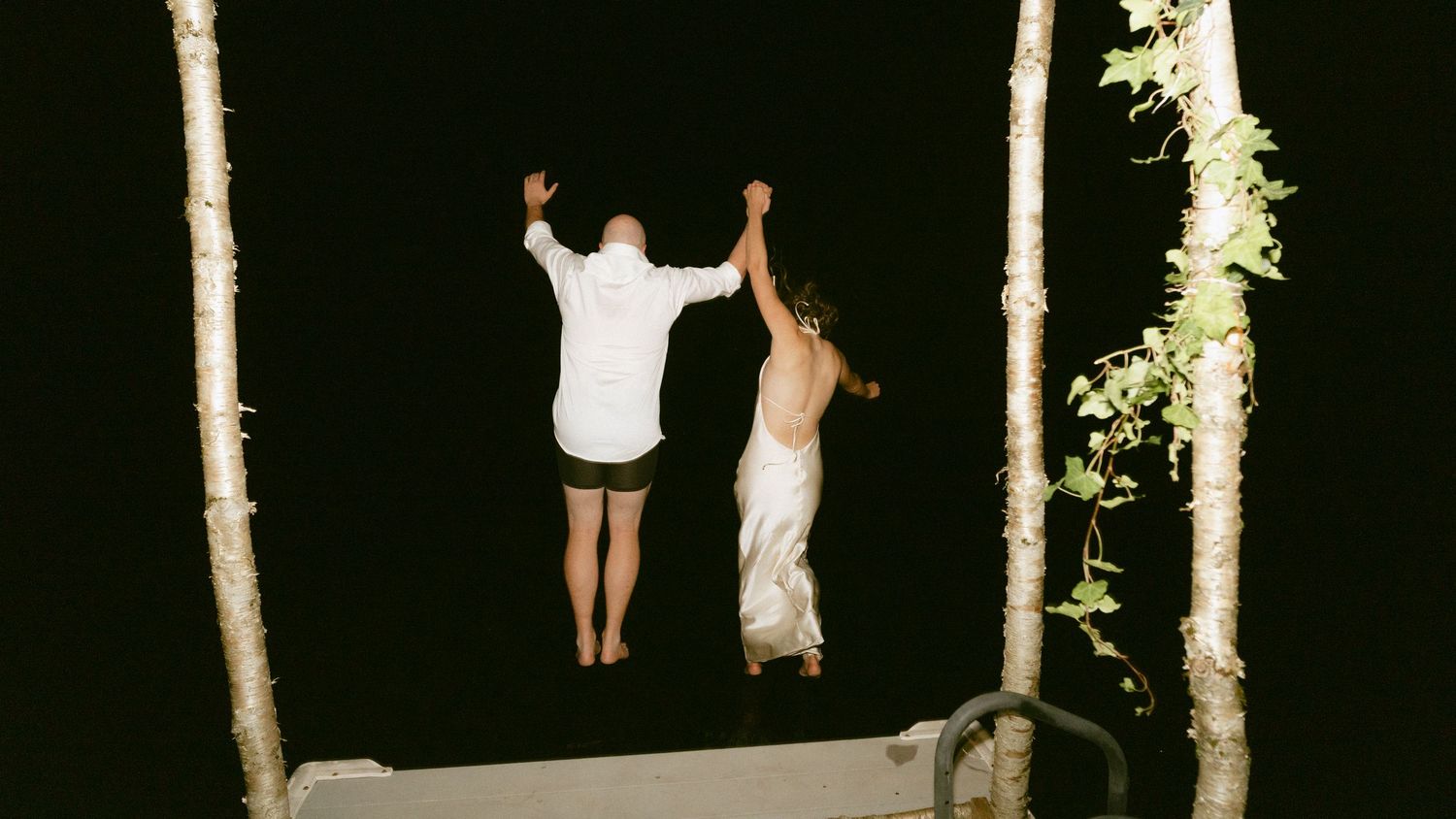 Bride and groom jumping hand-in-hand off a dock into dark water at night during wedding celebration.