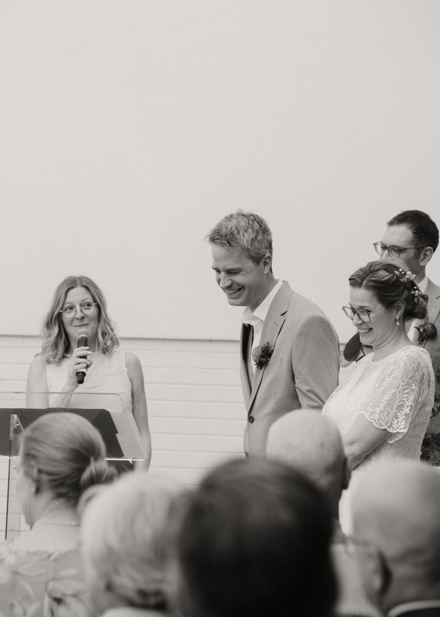 Black and white photo of smiling bride and groom during wedding ceremony with officiant reading from a podium.