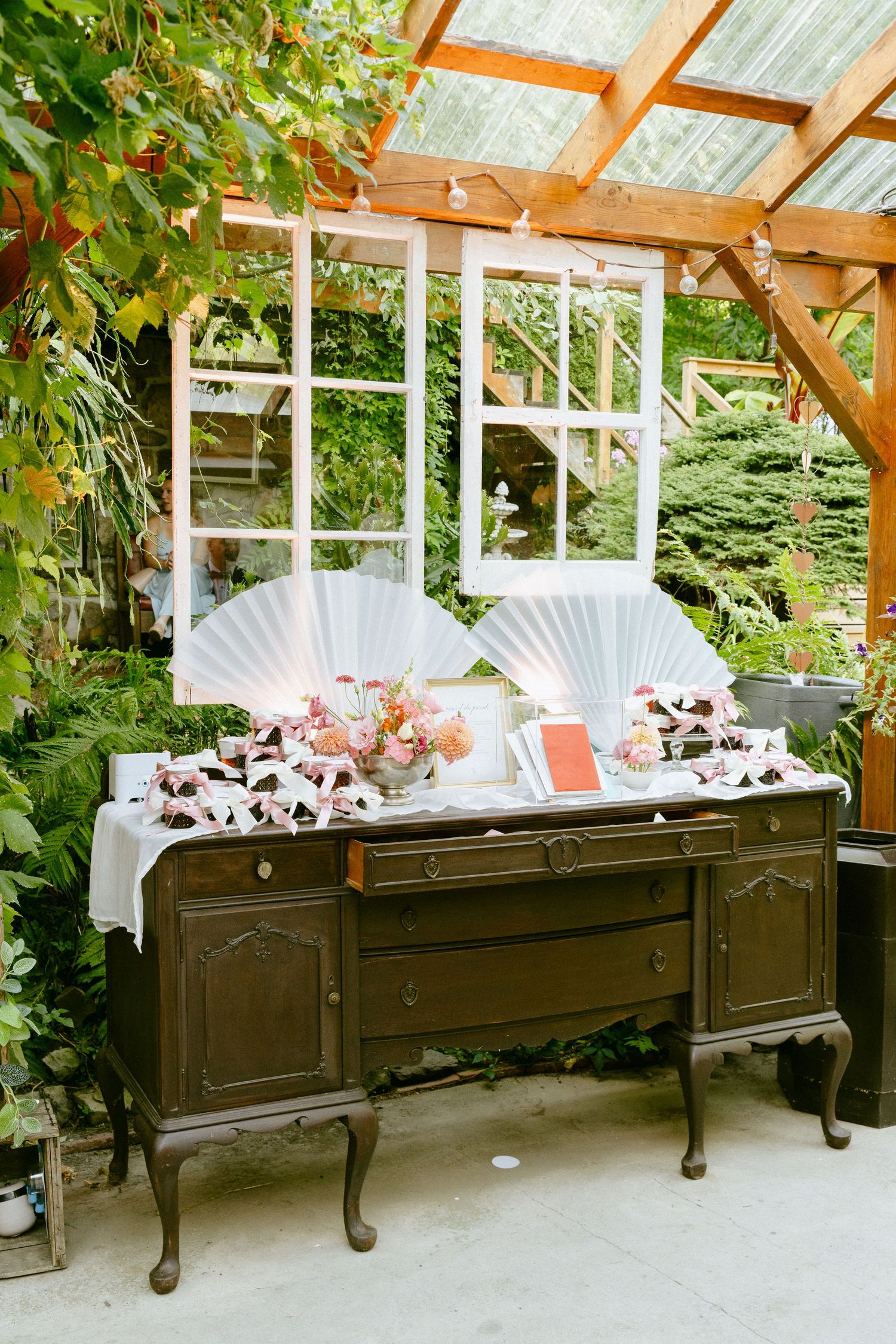 Vintage dark wood sideboard decorated with white fans, pink flowers, and wedding favors in a greenhouse.