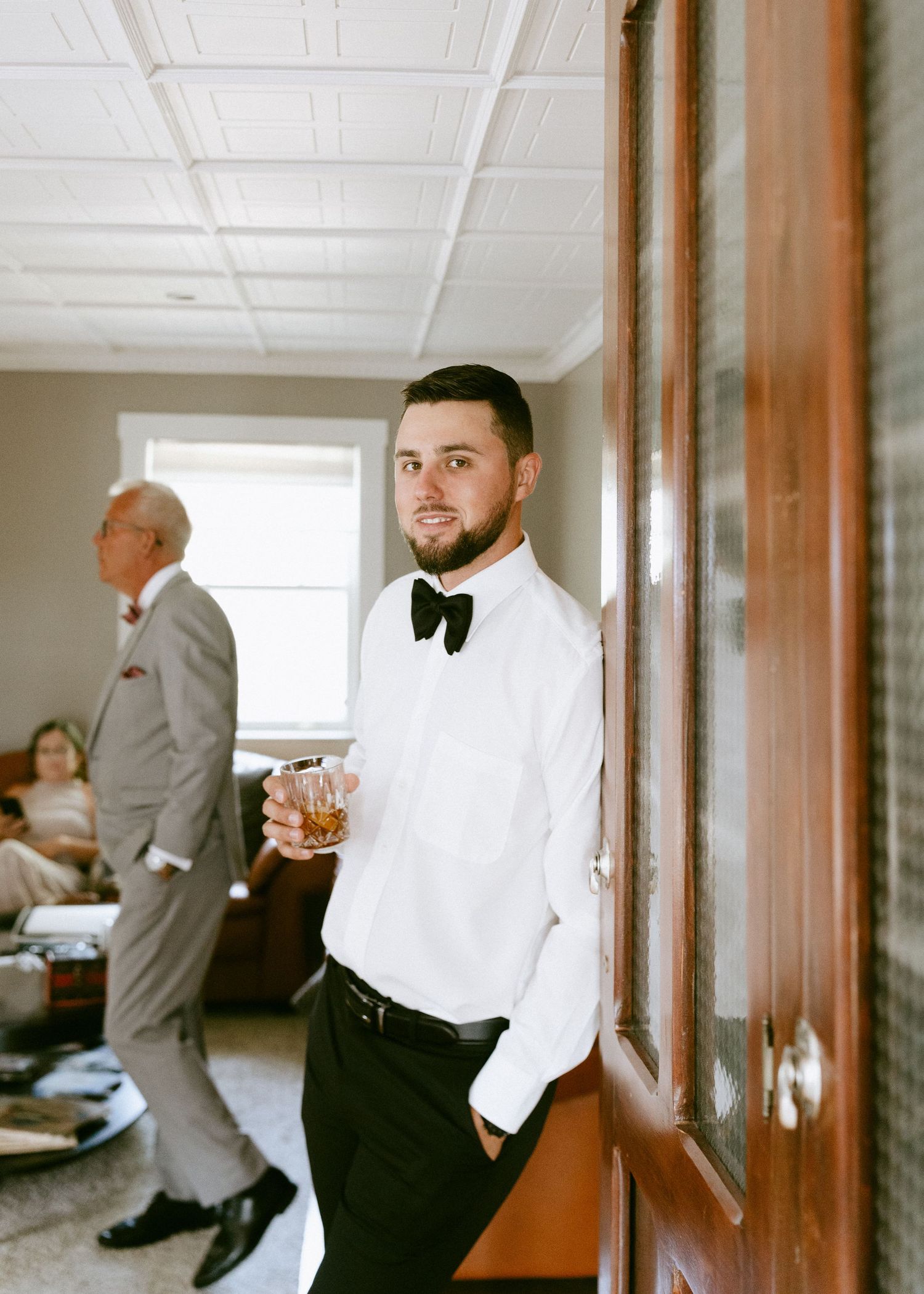 A groom in a white shirt and black bow tie holds a whiskey glass before his wedding.