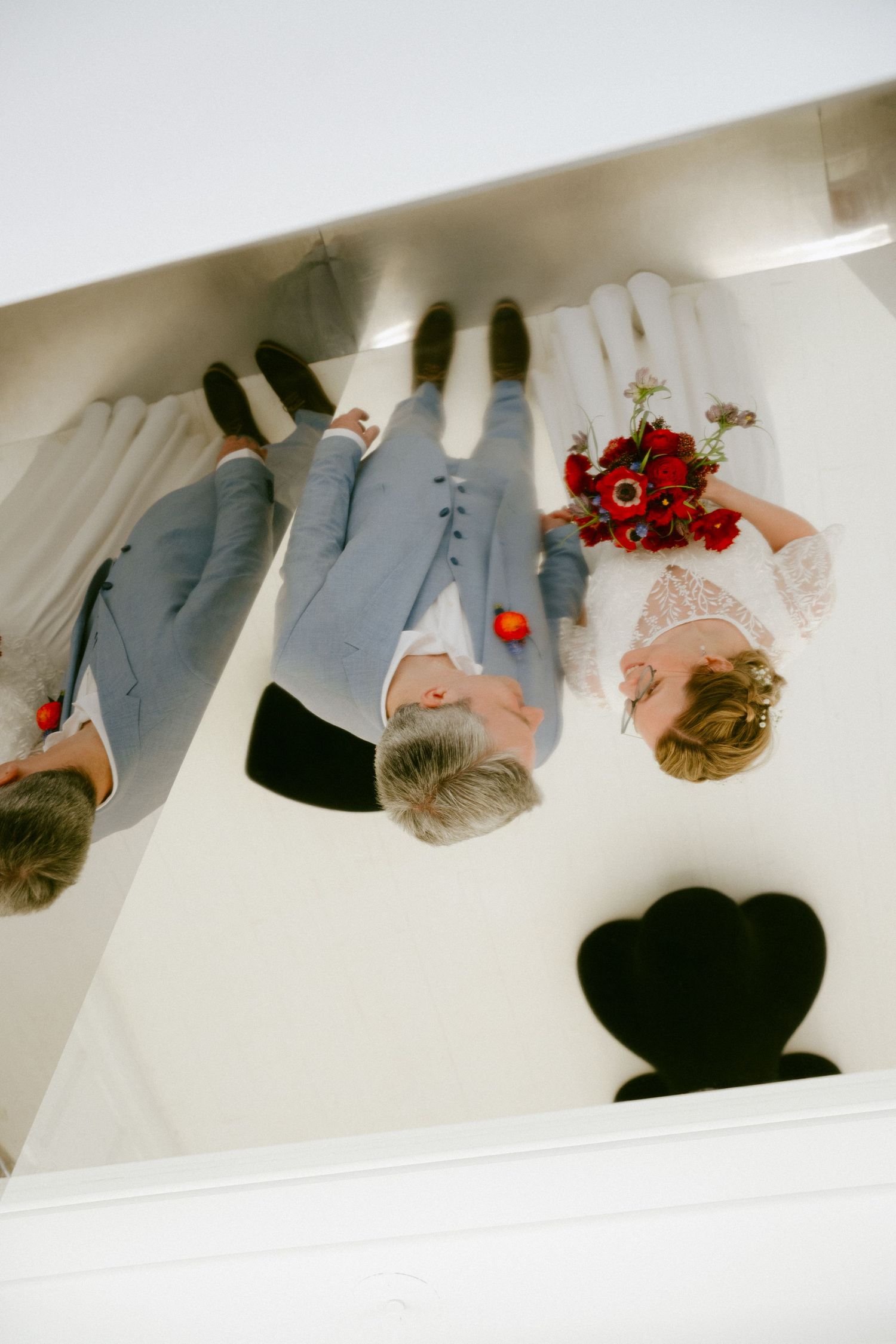 Aerial view of bride and groom in wedding attire holding red bouquet on white surface.