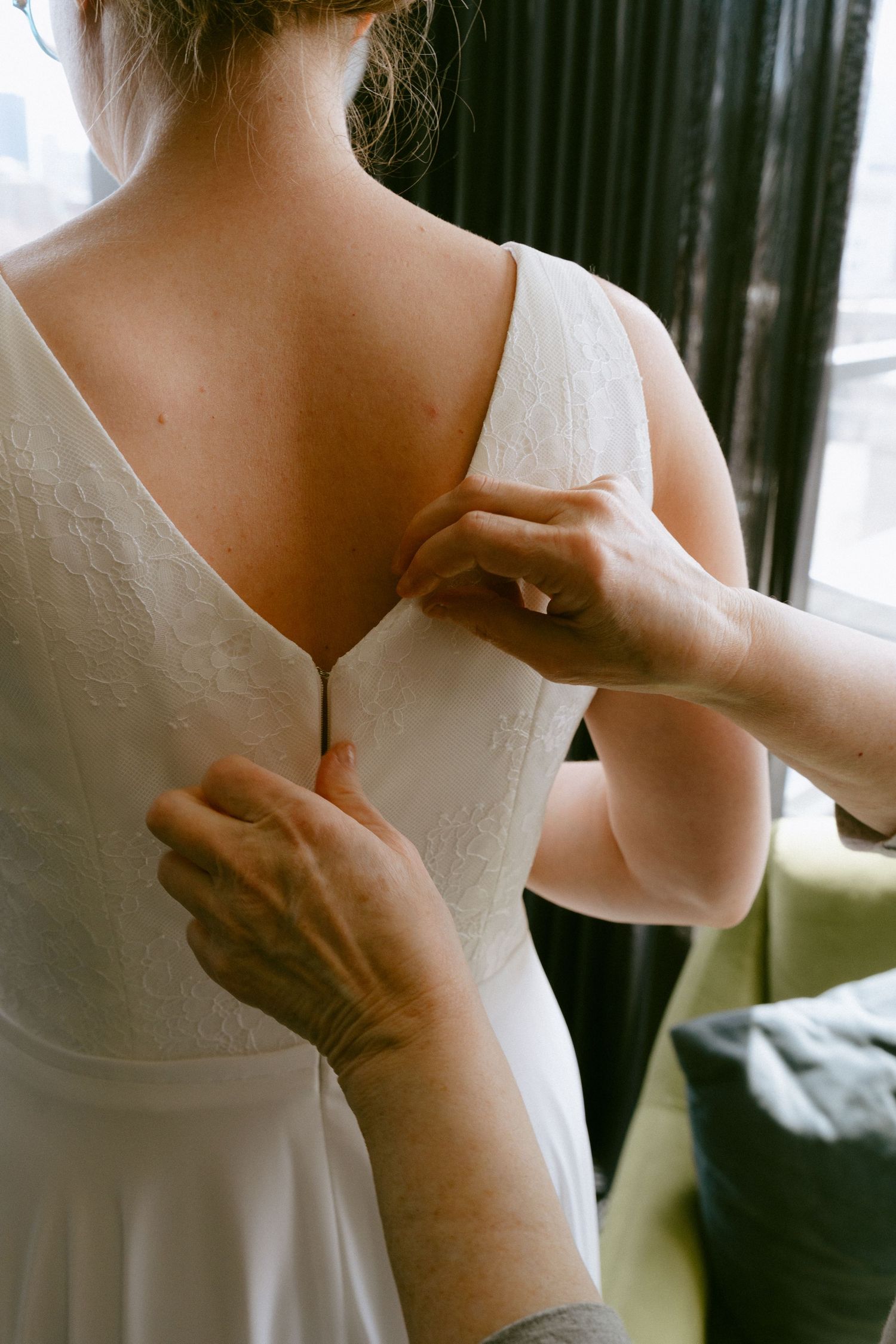 Assistant helps bride zip up elegant white lace wedding dress from behind near window.