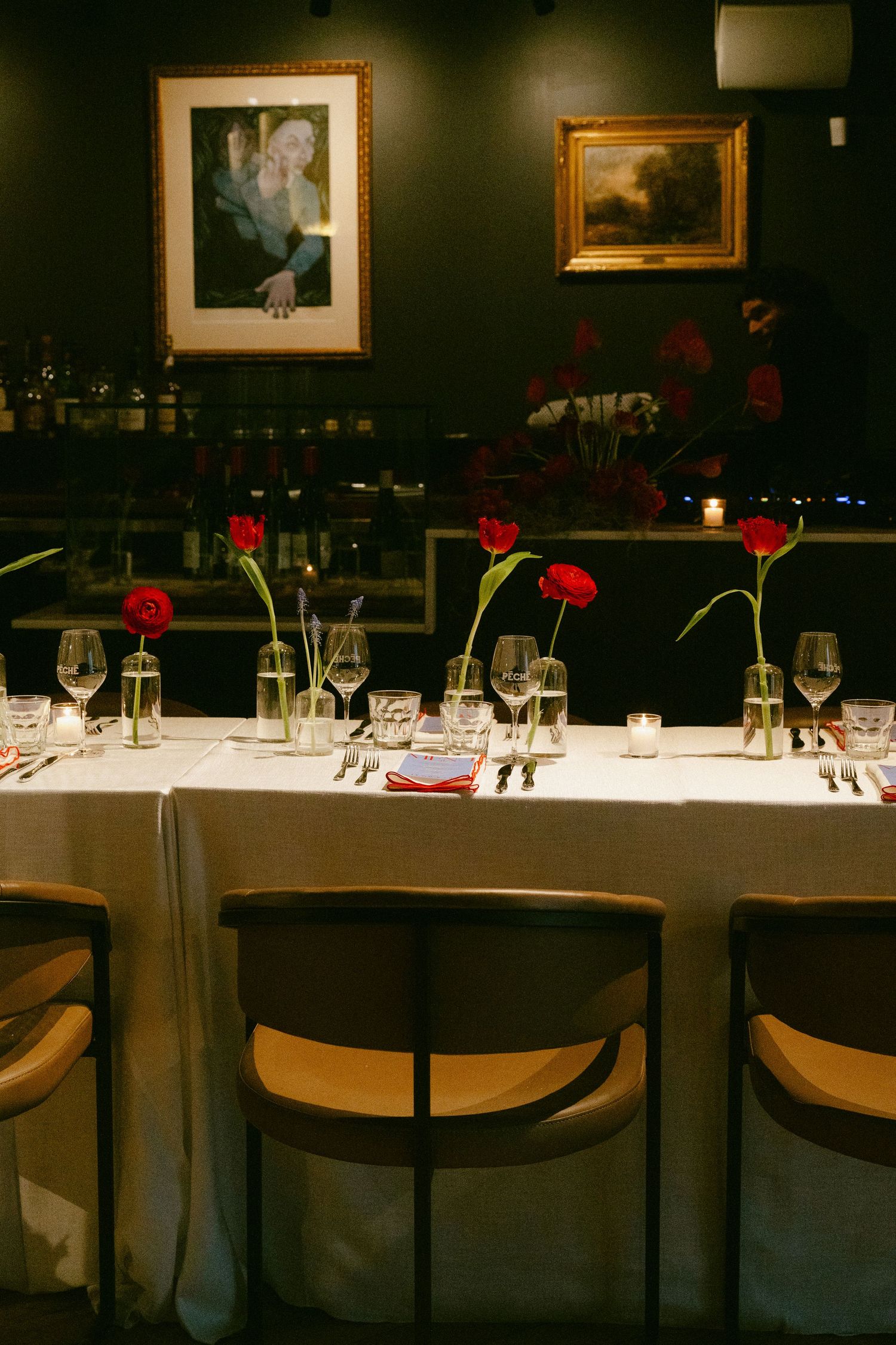 Intimate restaurant dinner table with single red tulip stems, wine glasses, and classic art paintings on dark walls.