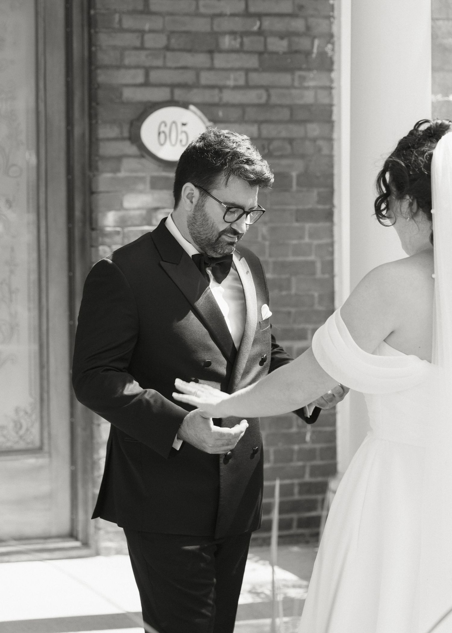 Groom in black tuxedo holds brides hand during first look at brick building entrance.