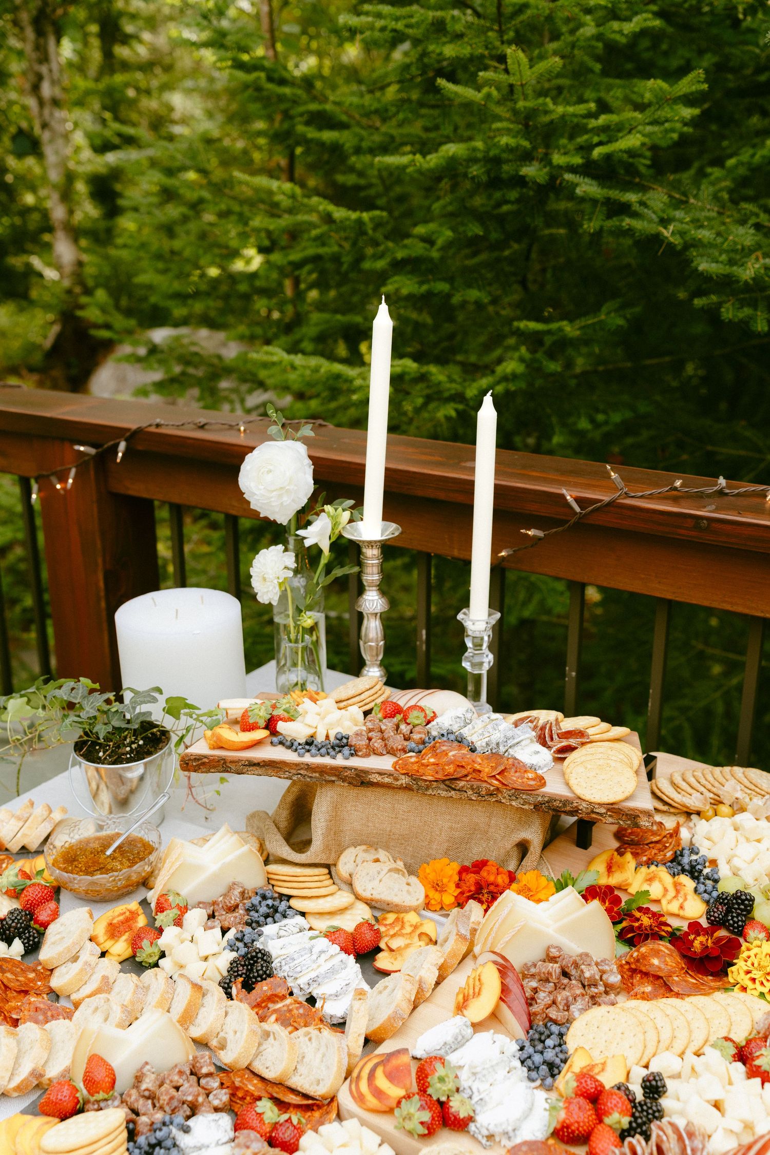 Outdoor wedding charcuterie table with candles, white roses, fruits, cheeses, and meats on a forest deck.
