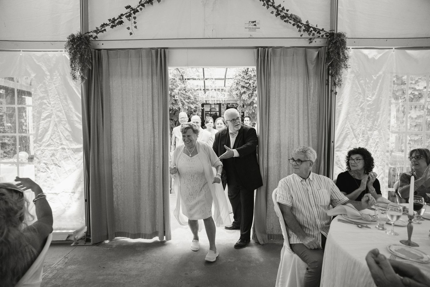 Black and white photo of bride and groom entering wedding reception tent greeted by seated guests.