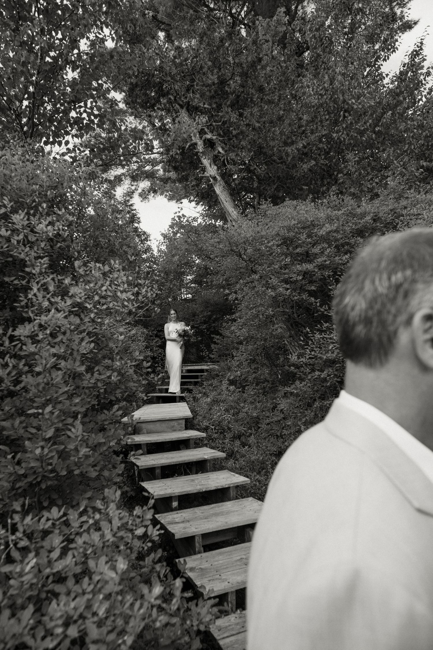 Black and white photo of a bride descending wooden stairs through lush garden greenery.