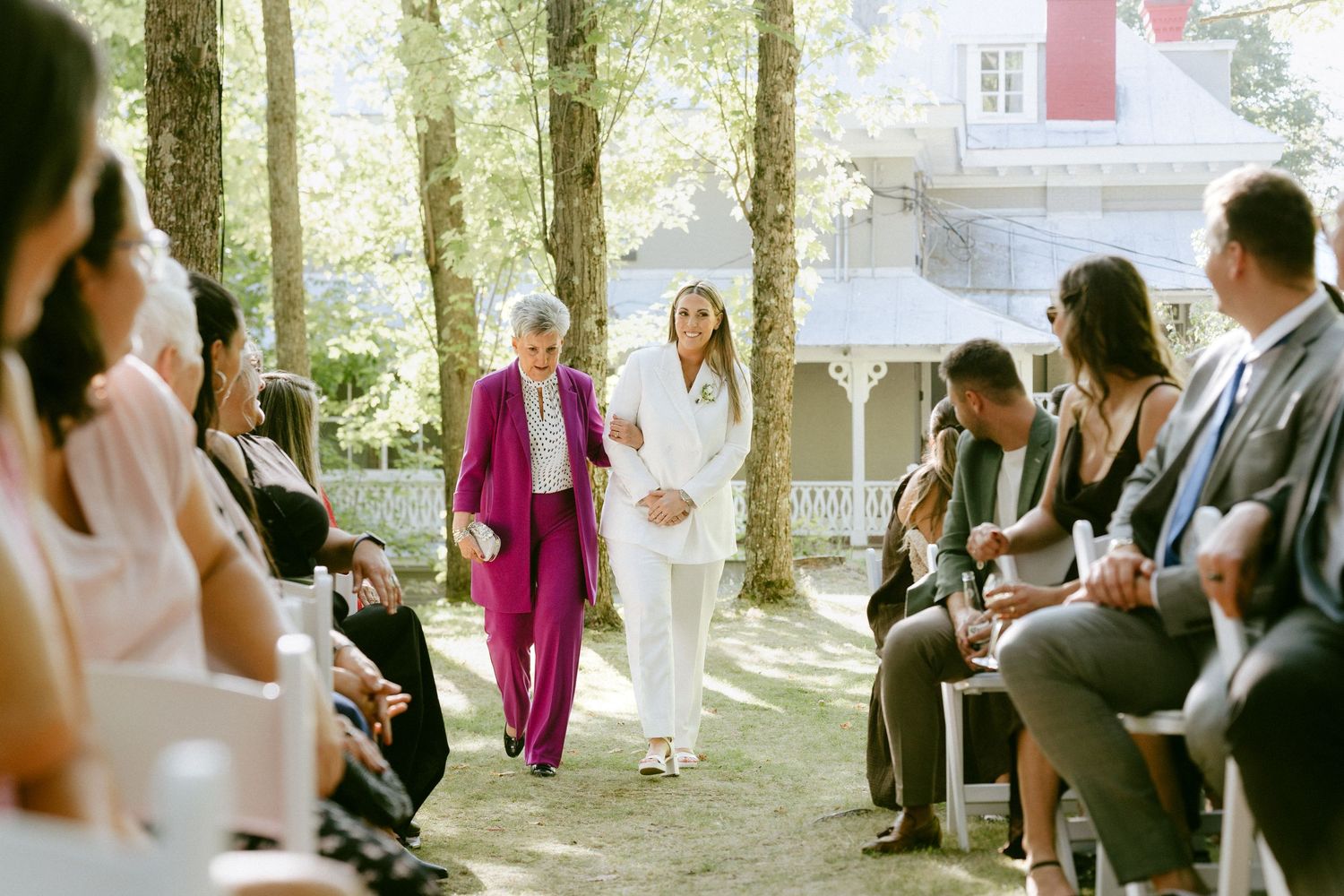 Bride in white suit walks down aisle with woman in purple suit at outdoor wedding.