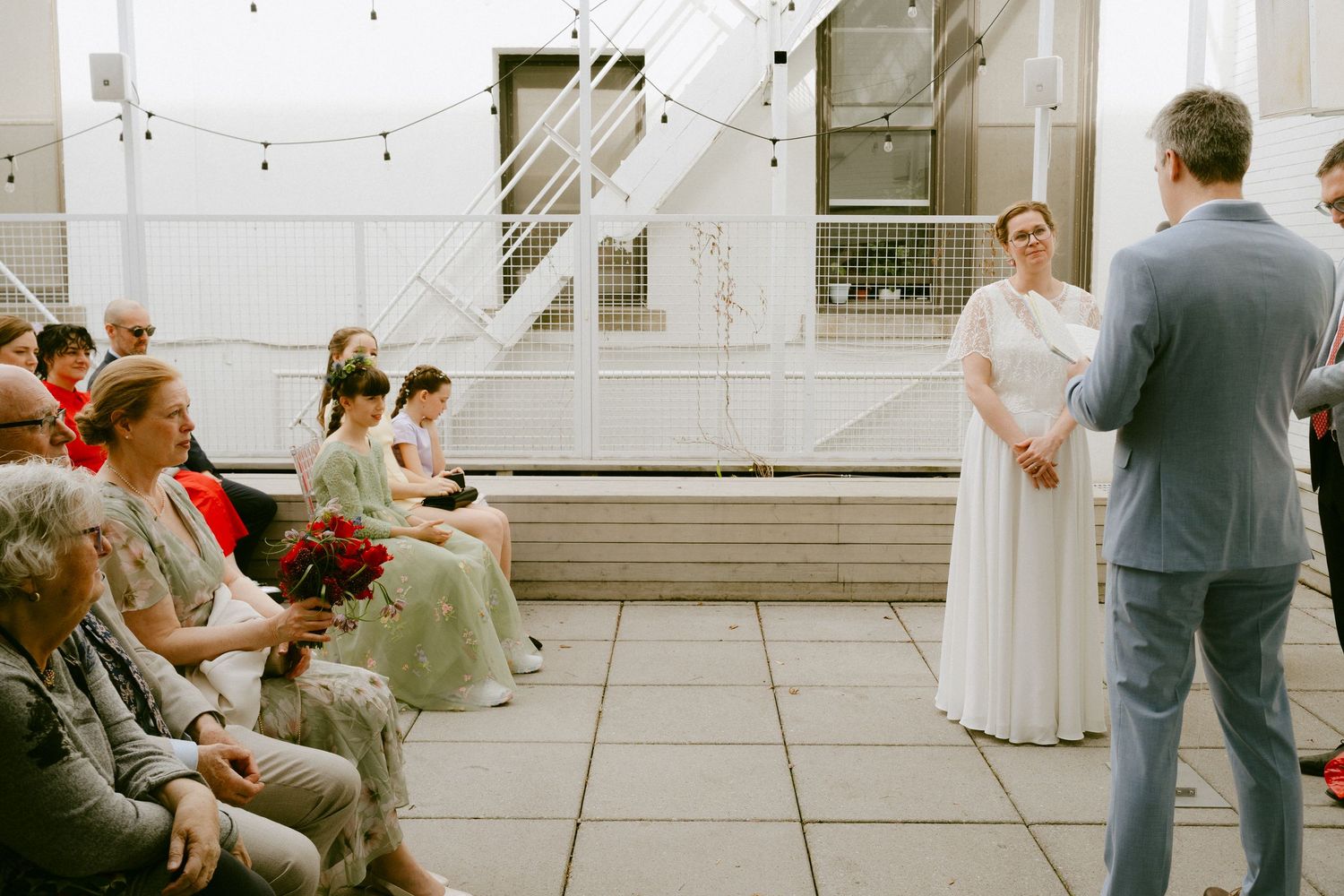 Bride in white gown stands facing groom during outdoor rooftop wedding ceremony with seated guests.