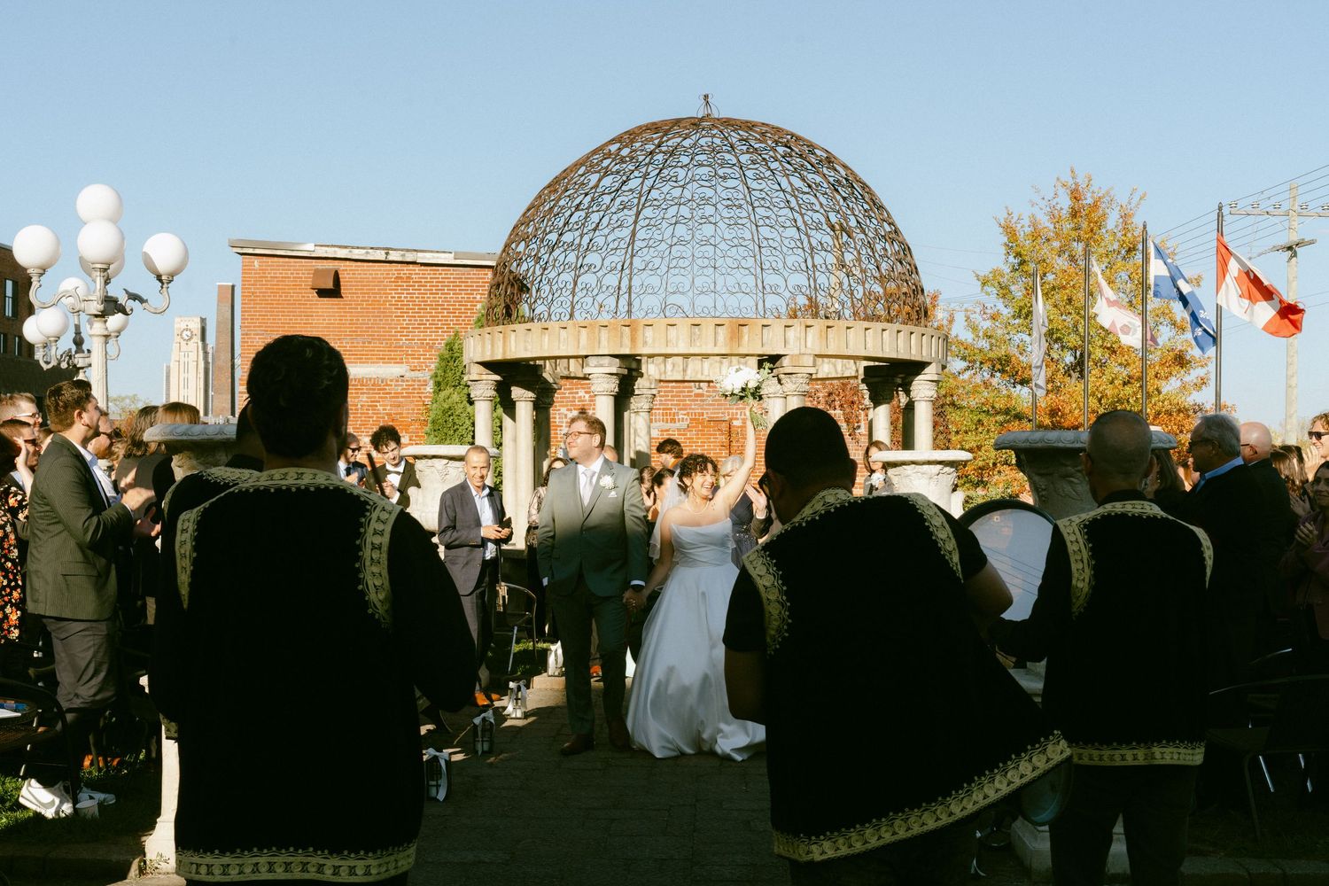 Wedding ceremony guests in traditional attire watch couple exchange vows before ornate glass dome gazebo.