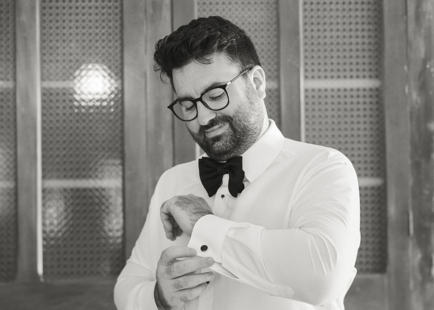 A groom in a white shirt and black bow tie adjusts his cufflinks in black and white.